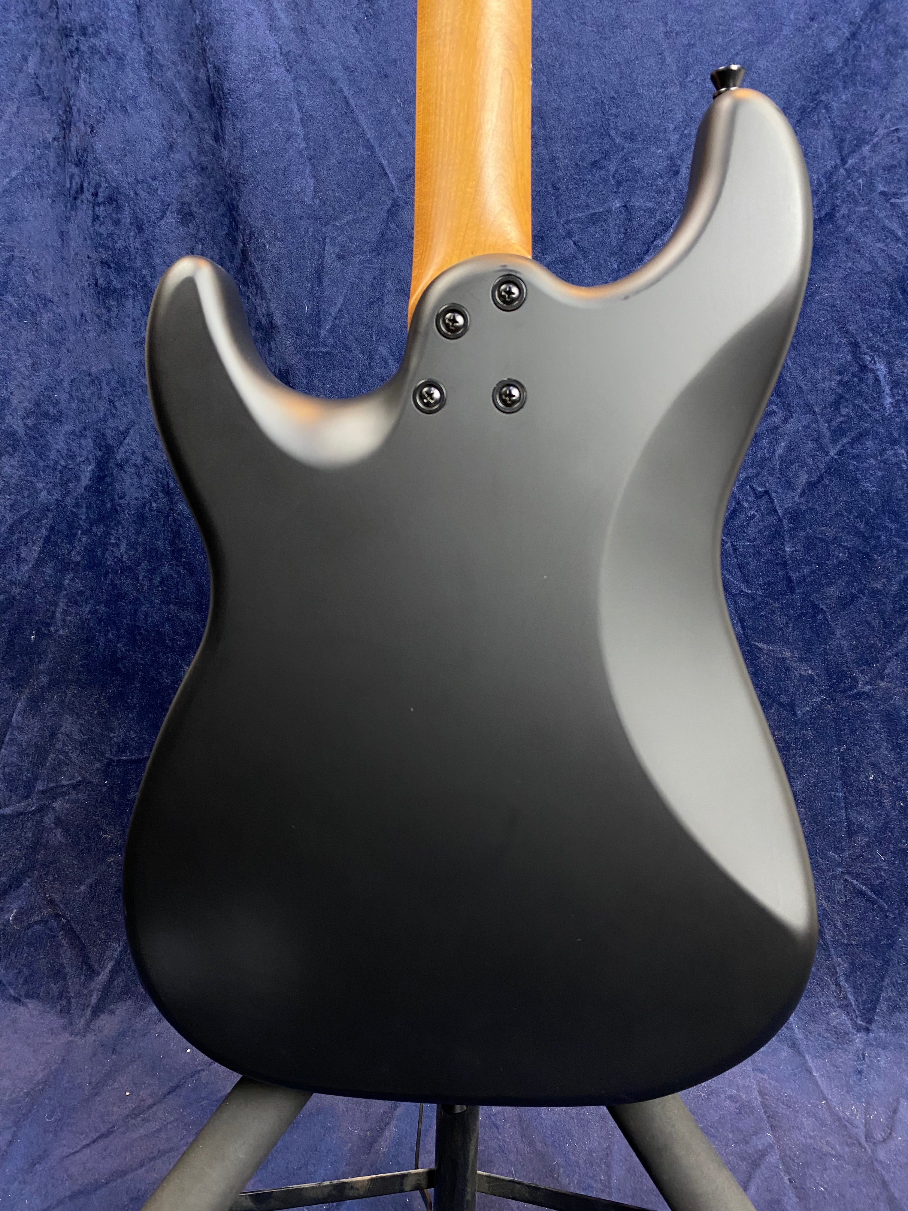 Matte black electric guitar body with a natural wood neck, four bolts attaching the neck, shown from the back against a dark blue background.