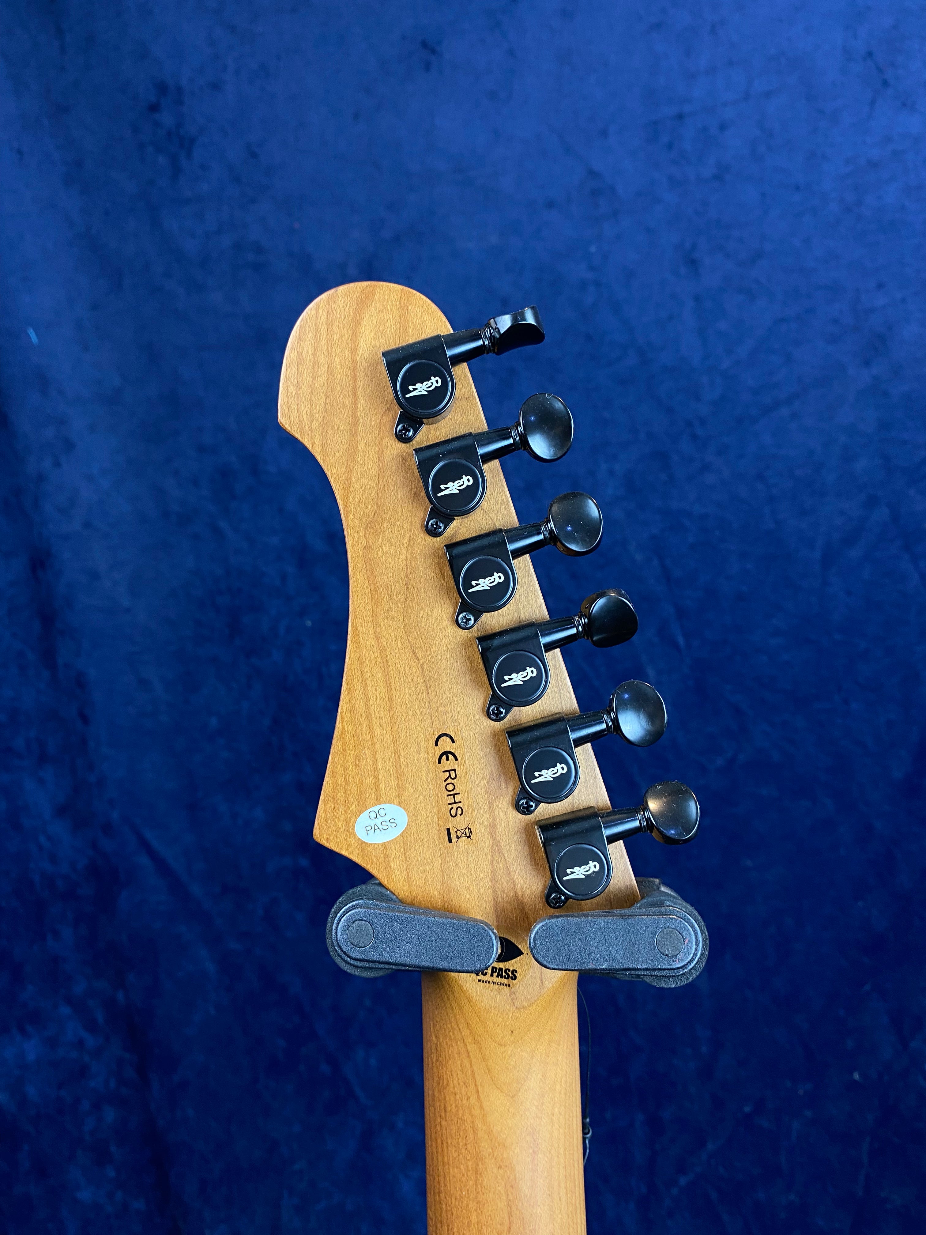 Six black tuning pegs on the back of a wooden electric guitar headstock, with inspection and CE/RoHS certification stickers visible.