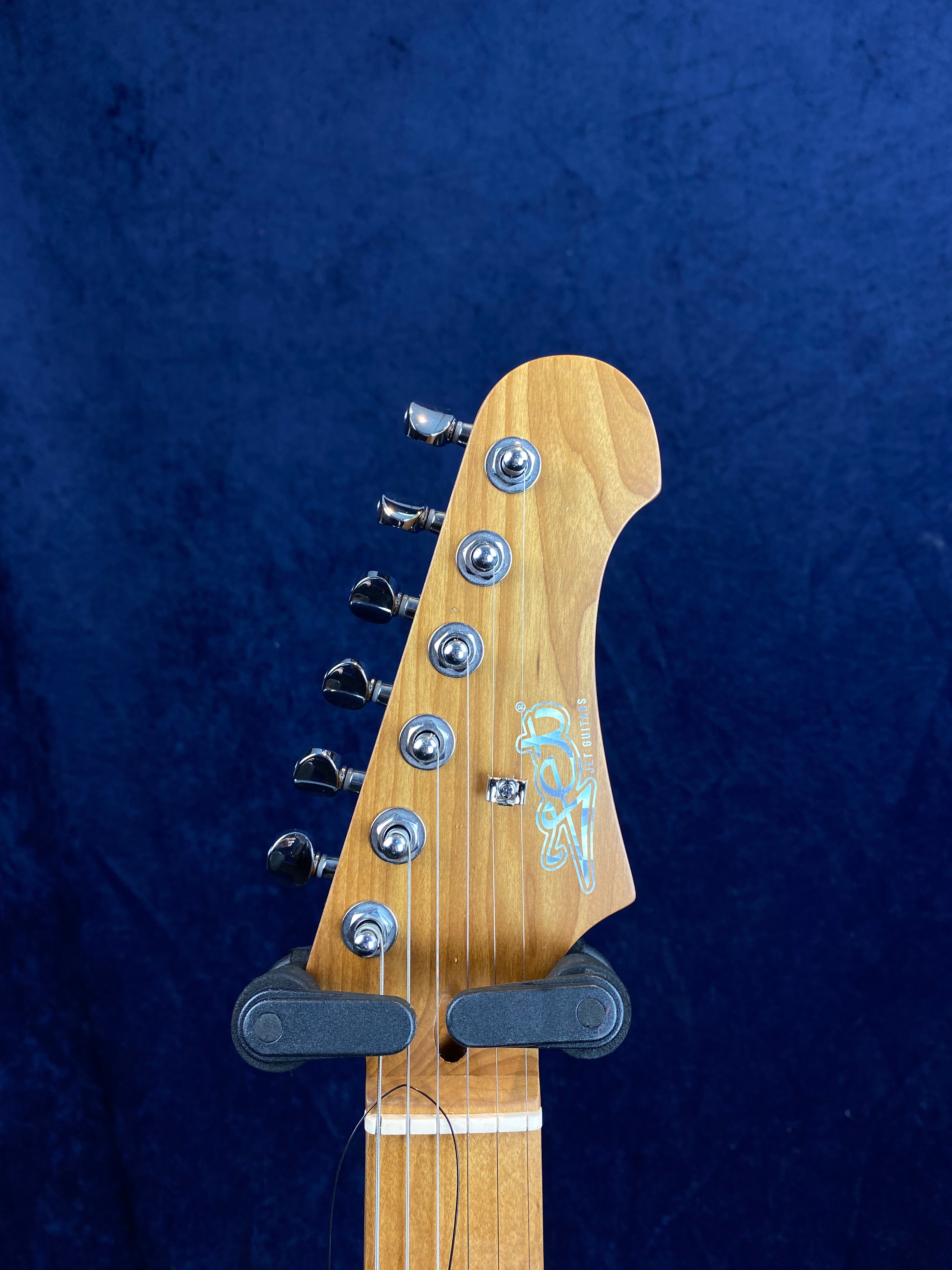 Tête de guitare électrique en bois clair avec six mécaniques chromées alignées et un logo stylisé sur fond bleu uni.