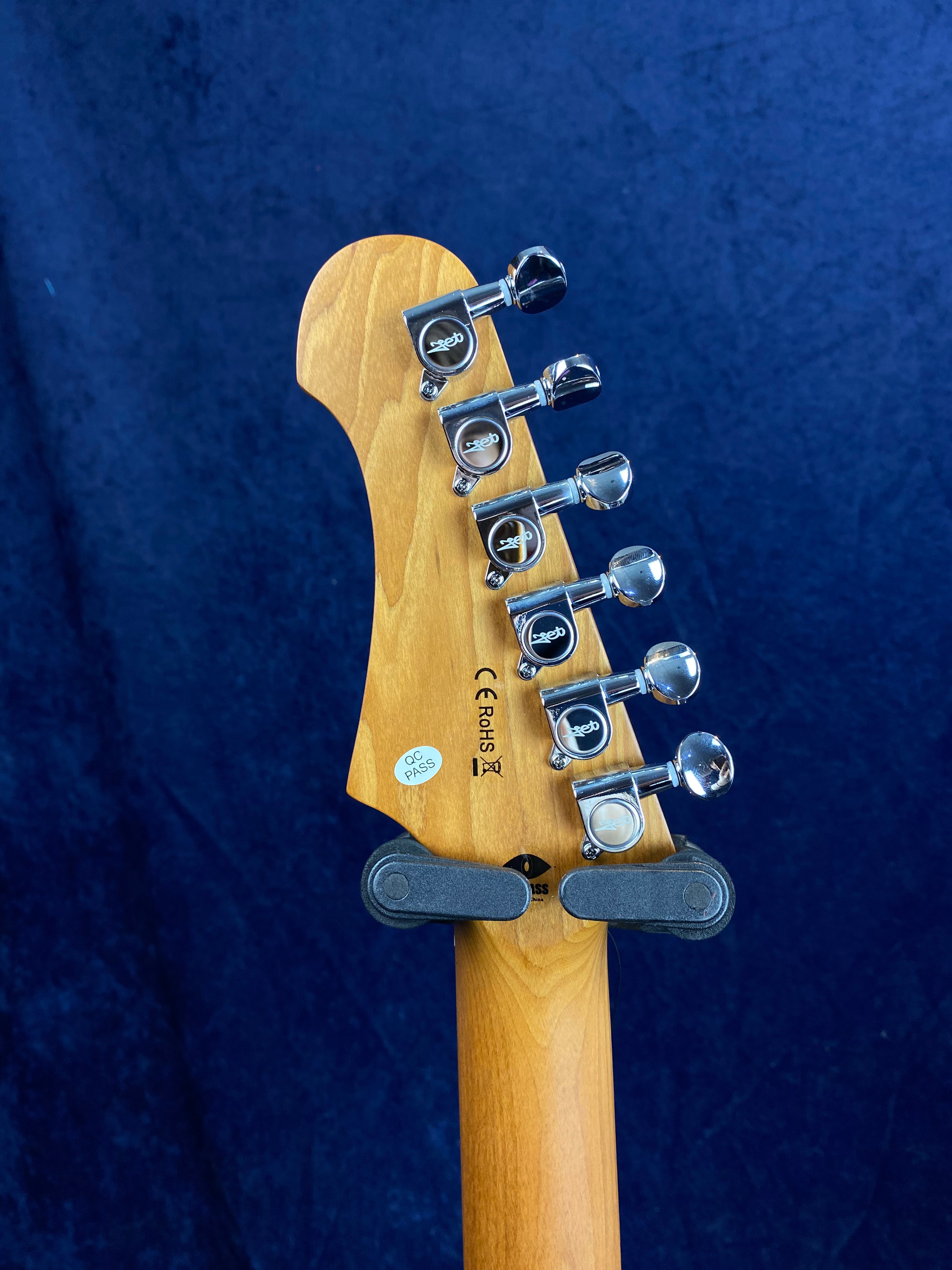 Tête de guitare électrique en bois clair avec six mécaniques chromées alignées, fond bleu foncé, autocollants de contrôle qualité visibles.