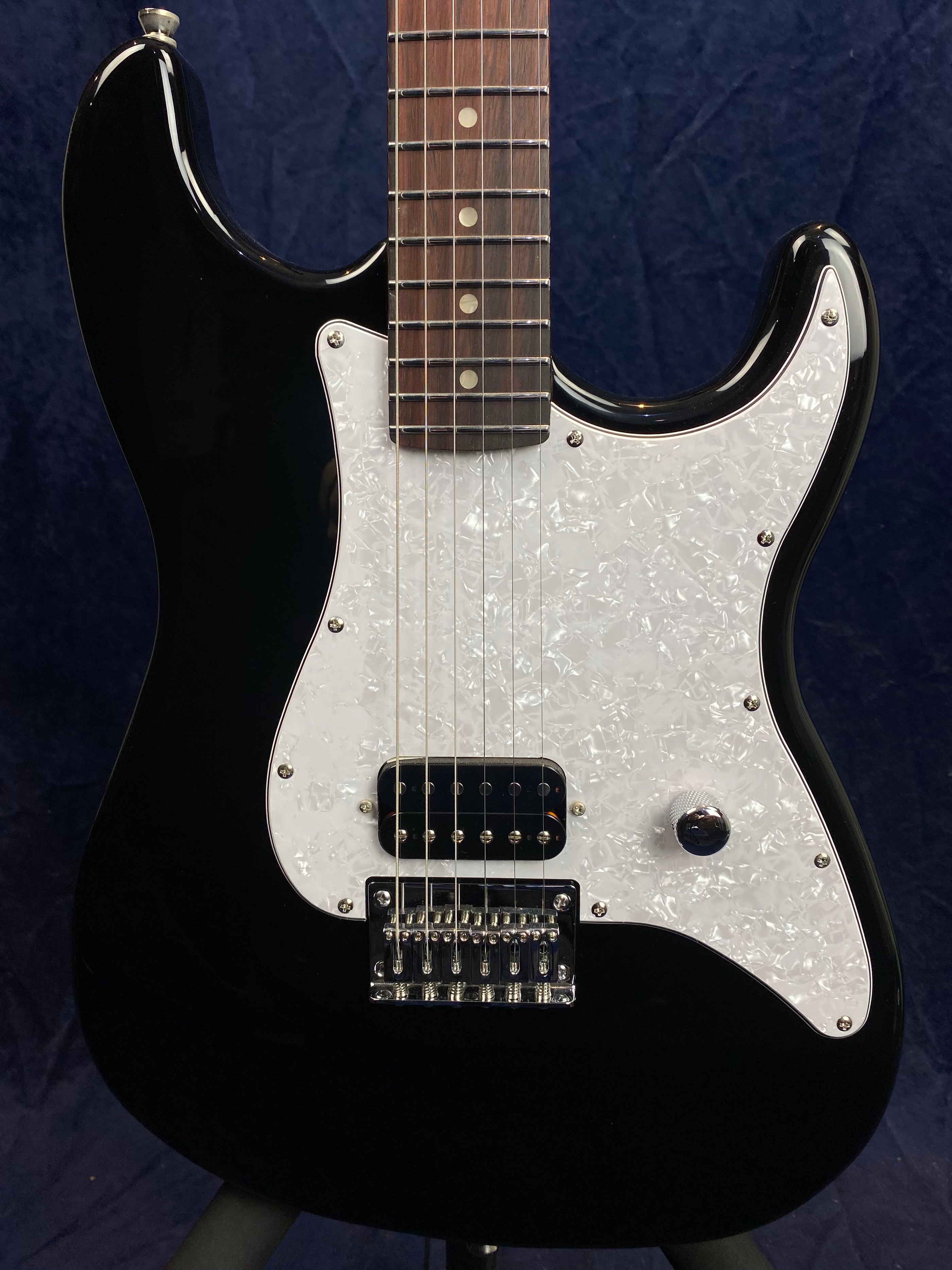 Black electric guitar with a white pearloid pickguard, one humbucker pickup, one control knob, and a chrome bridge.