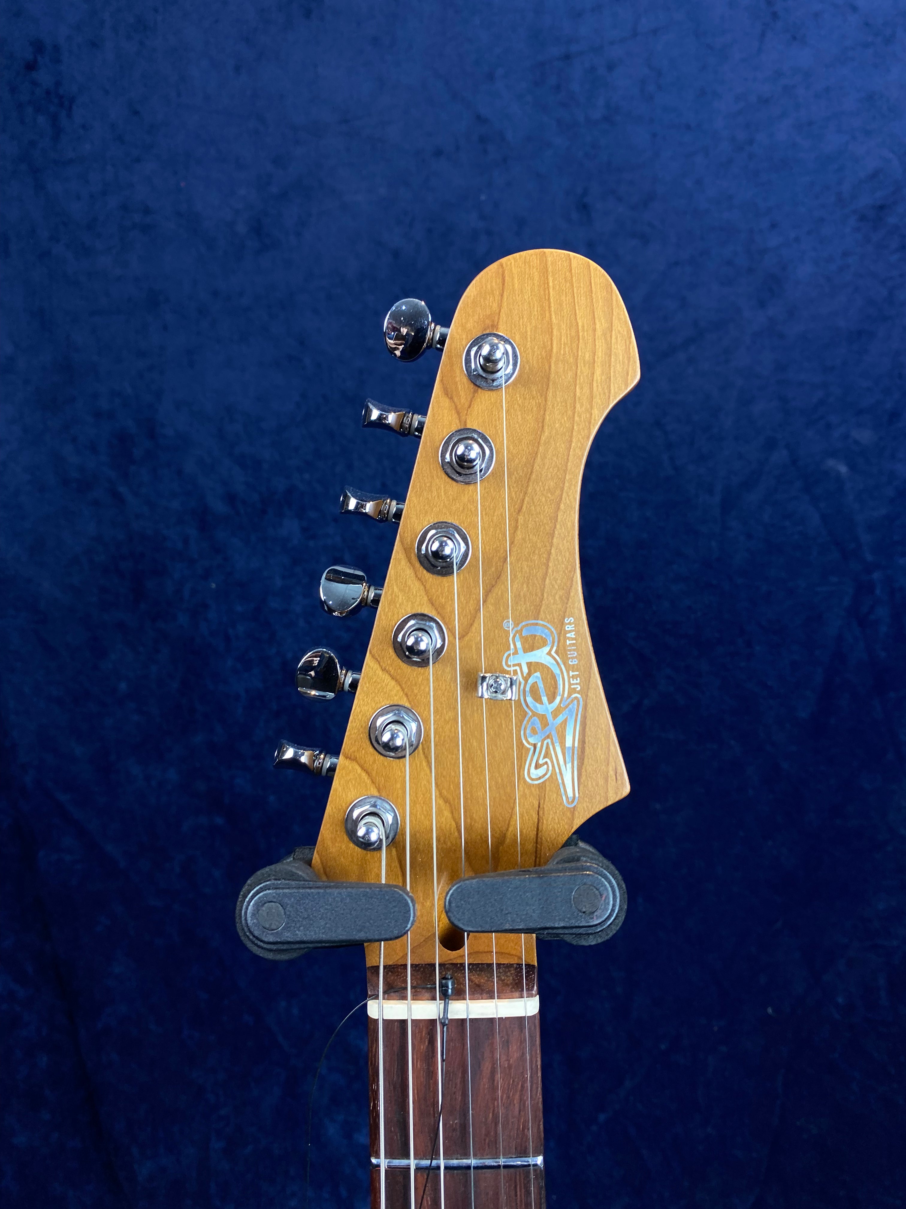 Light wood electric guitar headstock with six chrome tuning pegs and "JET GUITARS" logo, against a dark blue background.