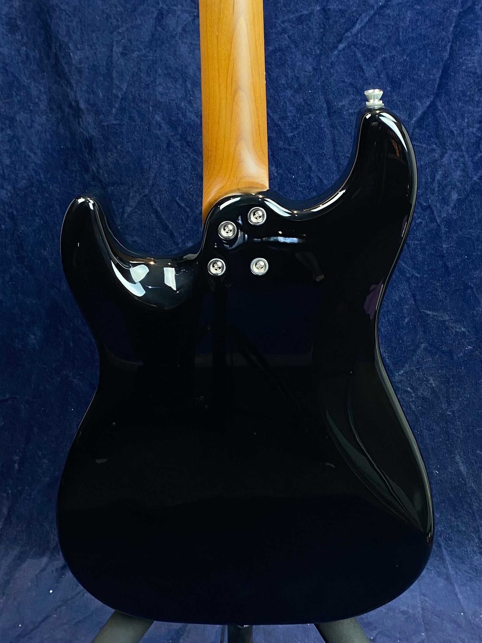 Glossy black electric guitar back with a natural wood neck, four neck bolts, and a metal strap button near the upper horn, set against a dark background.