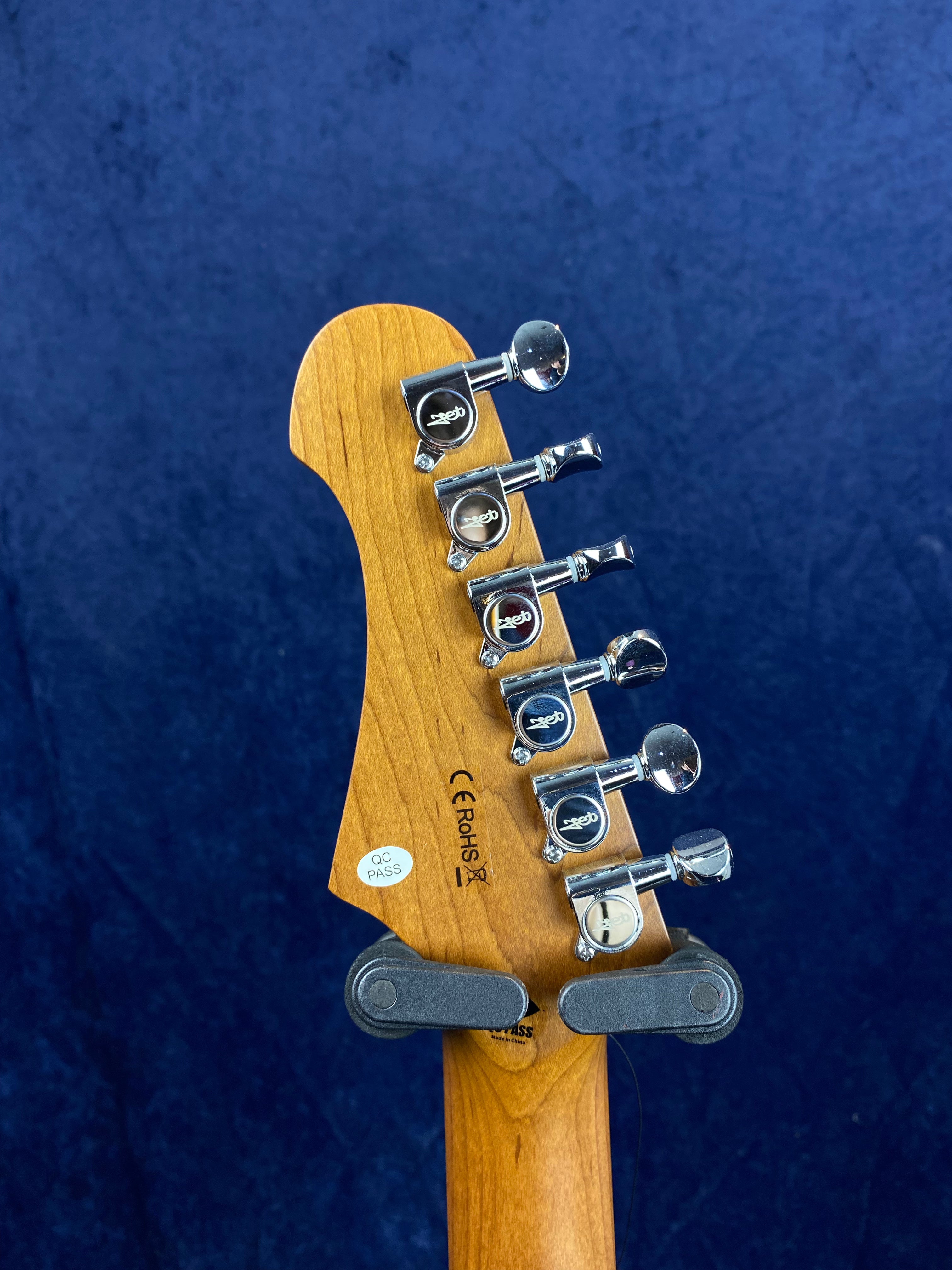 Six chrome tuners and QC PASS sticker on the back of a light wood electric guitar headstock, blue textured background.