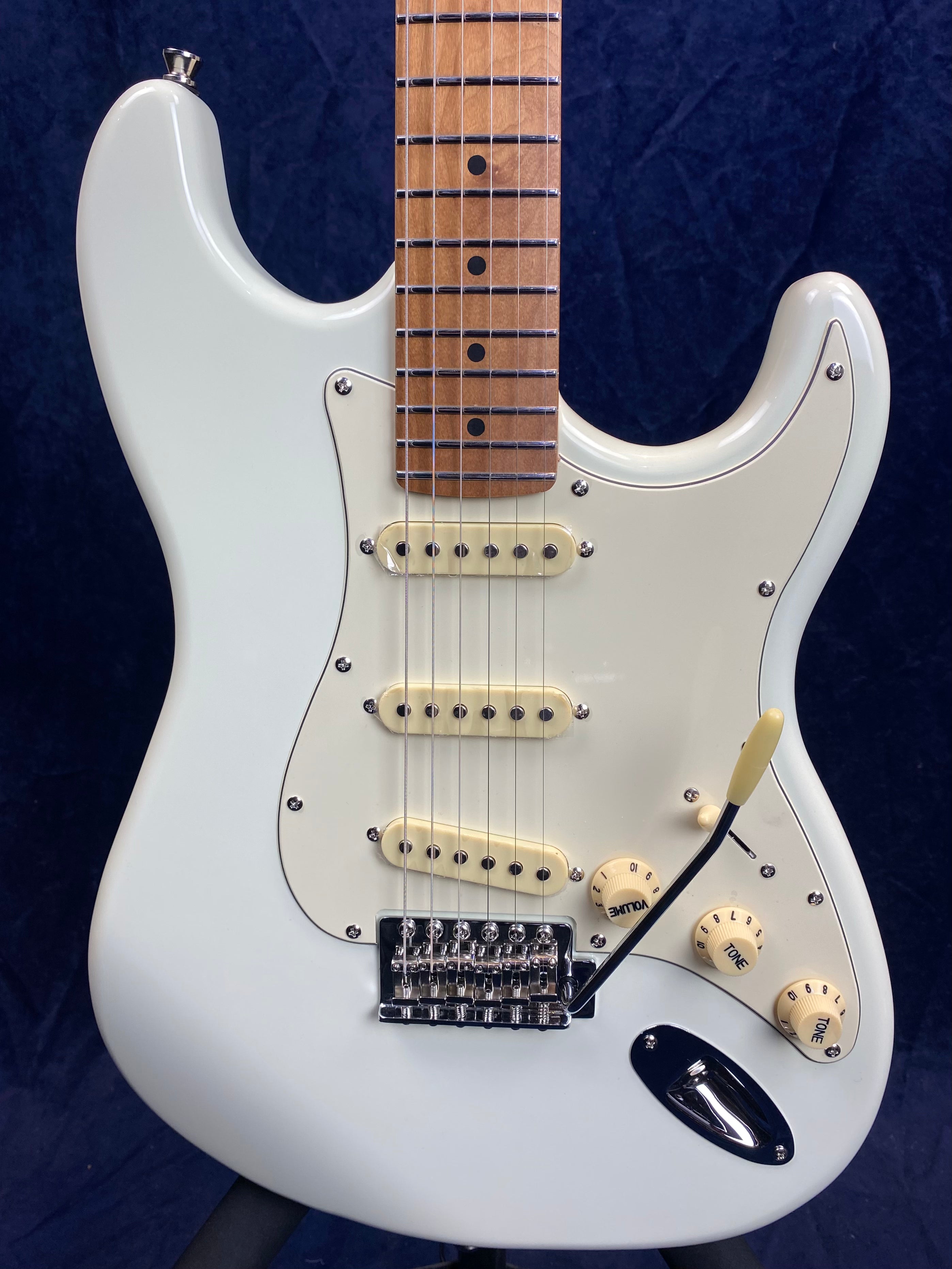 White electric guitar with three single-coil pickups, cream knobs, and a maple fretboard. The body has a curved shape and a vintage-style tremolo bridge.