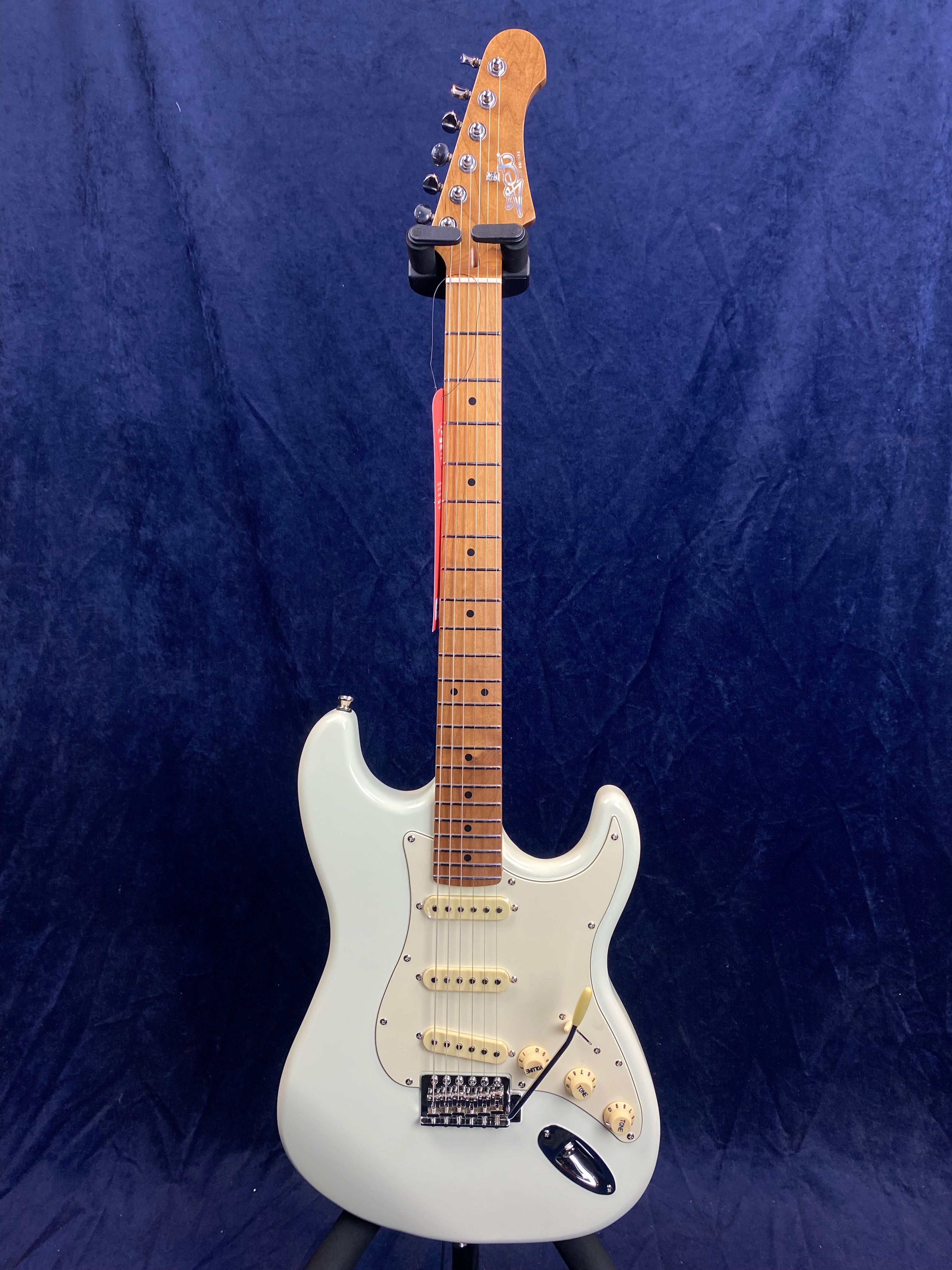 White electric guitar with three single-coil pickups, maple fretboard, and light-colored knobs, displayed upright against a dark blue background.