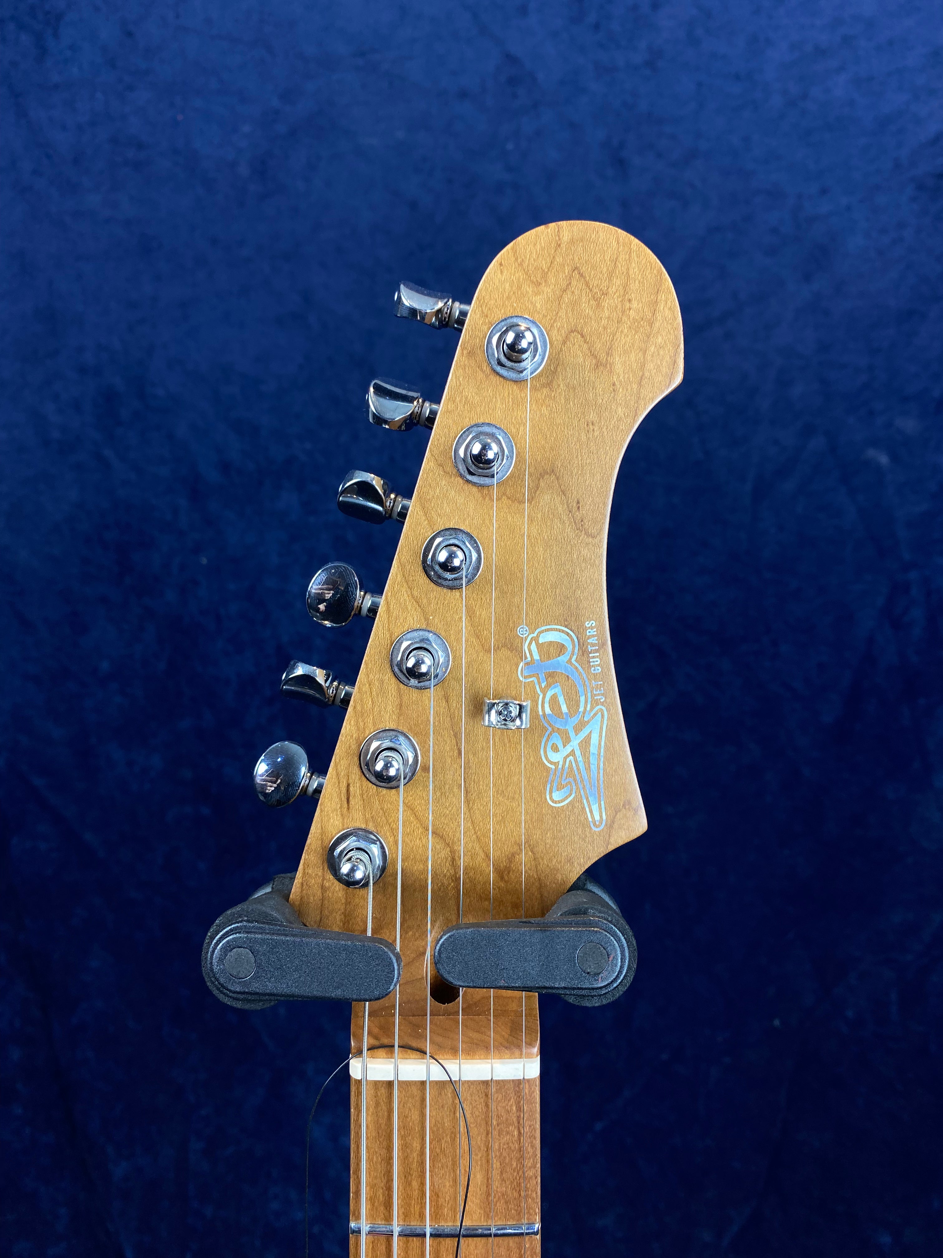 Light wood electric guitar headstock with six silver tuning pegs, string retainer bar, and a stylized white logo on a dark blue background.