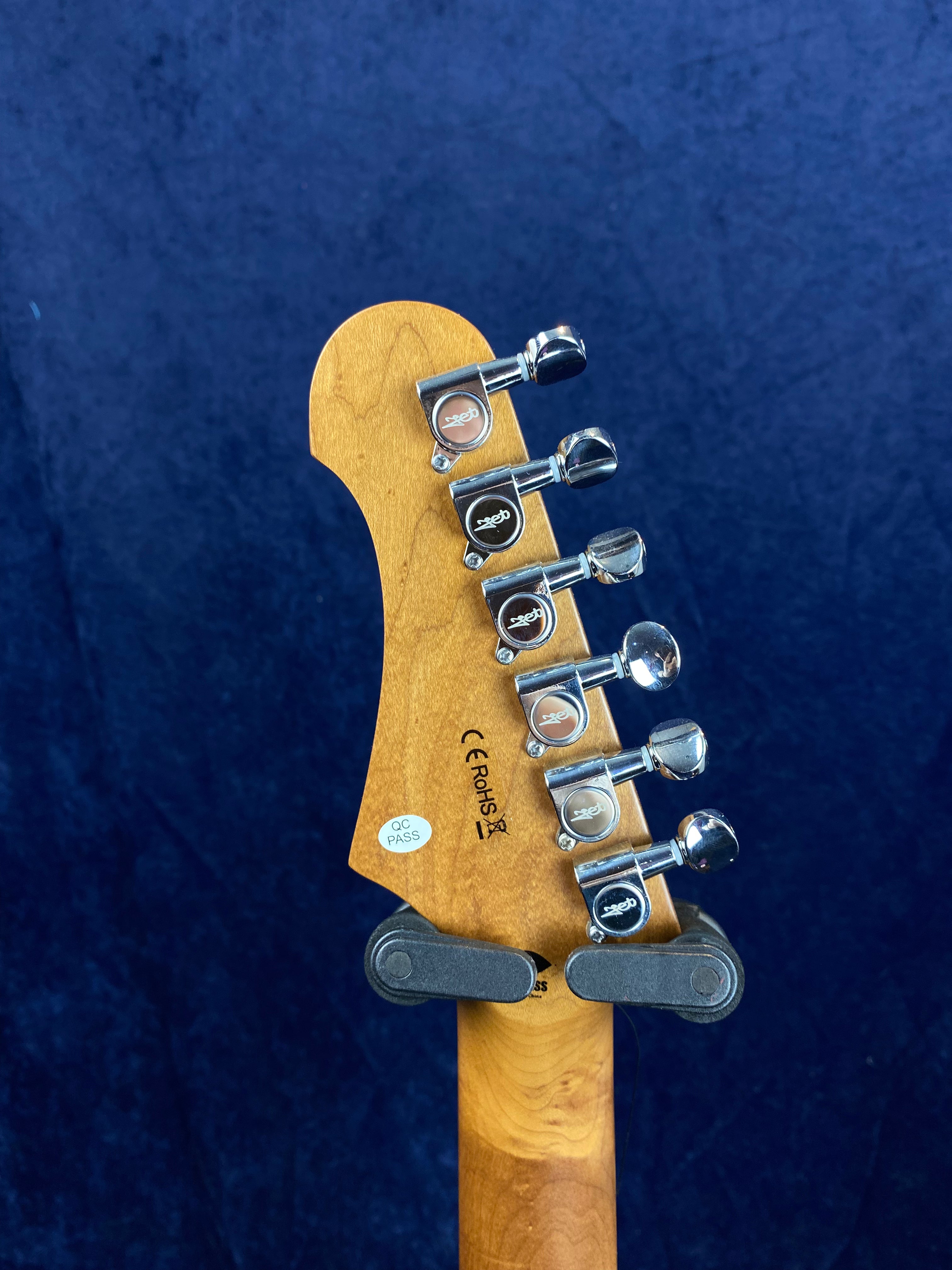 Close-up of an electric guitar headstock showing six chrome tuning pegs, certification stickers, and a wooden finish against a dark blue background.