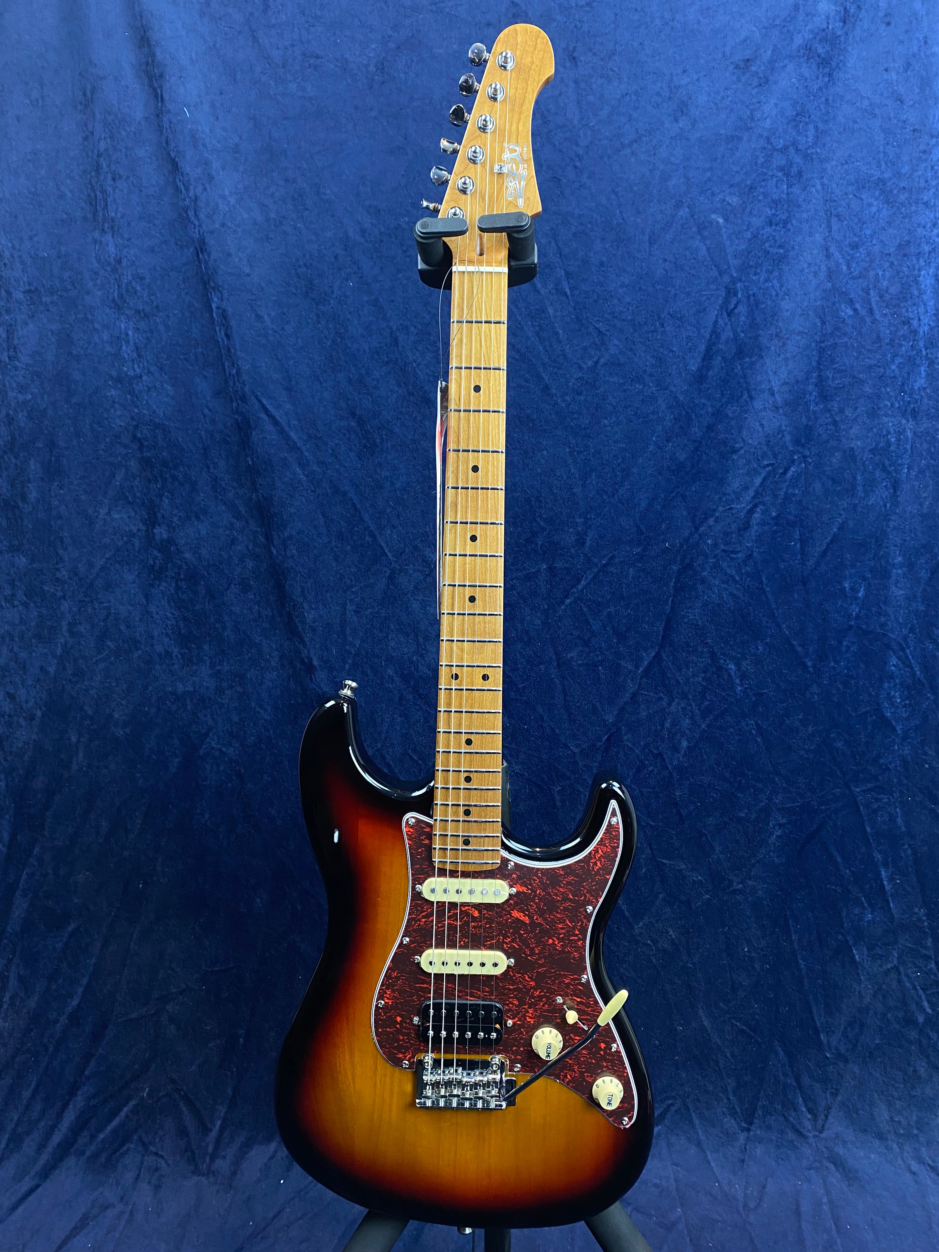Sunburst electric guitar with a maple neck, three pickups, tortoiseshell pickguard, and two control knobs against a dark blue background.