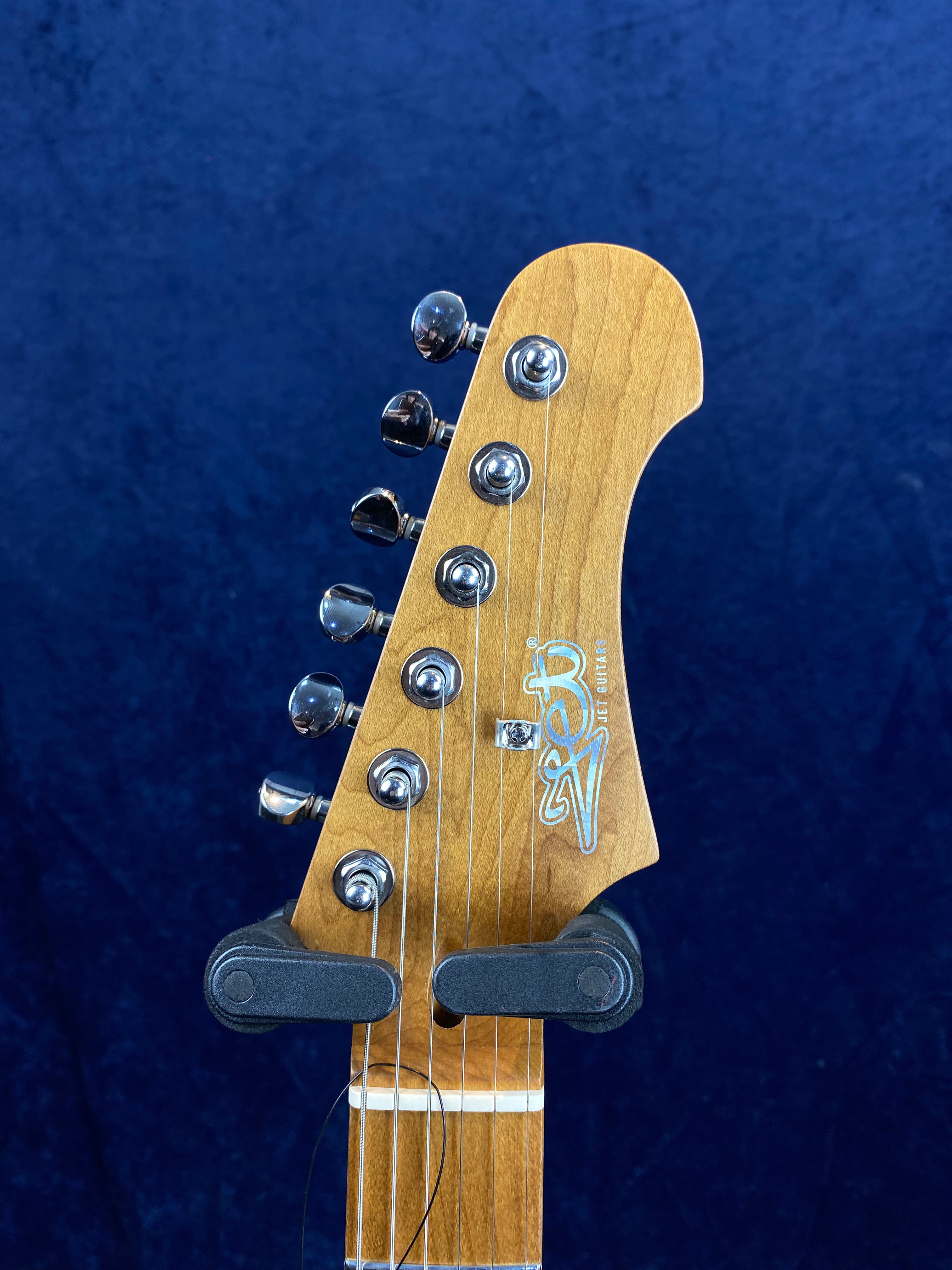 Natural wood electric guitar headstock with six chrome tuning pegs and the Jet Guitars logo, set against a dark blue background.
