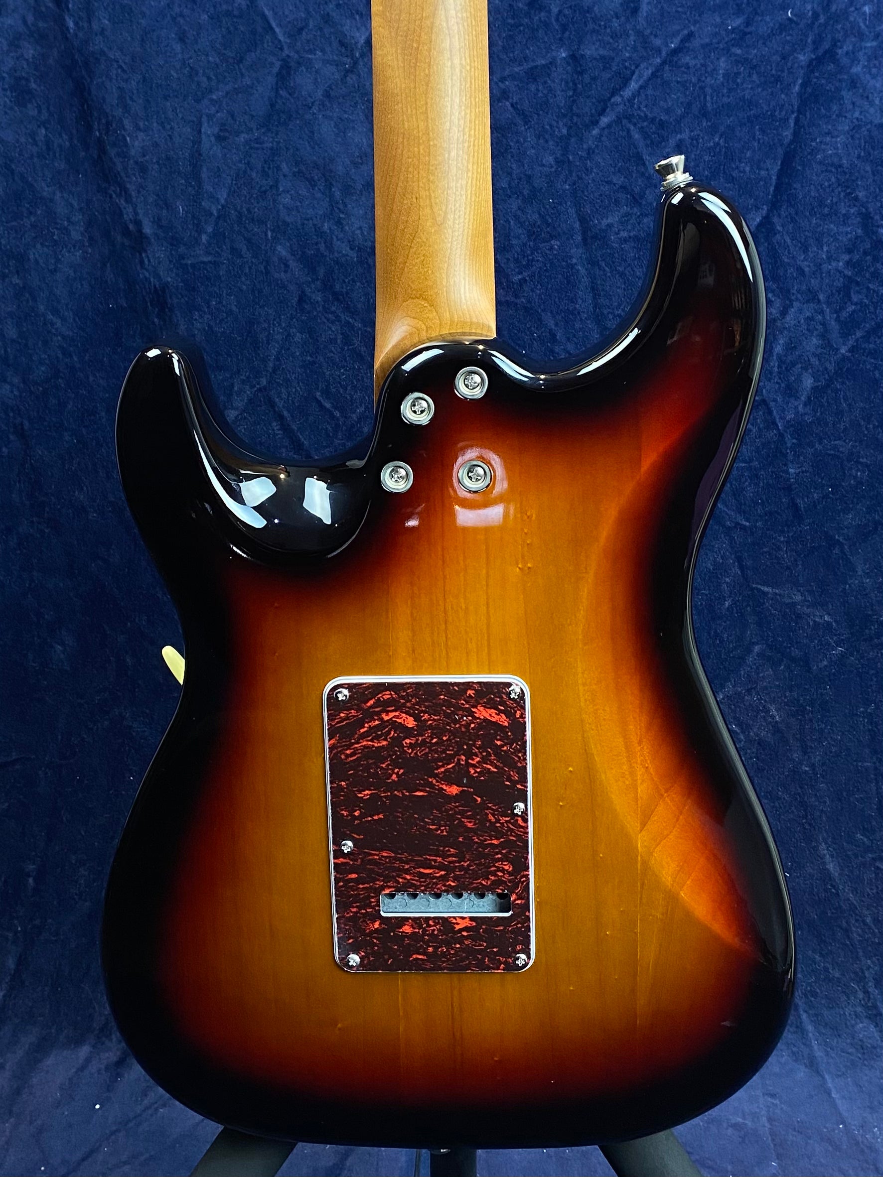 The back of a sunburst electric guitar with a natural wood neck and a red marbled tremolo cavity cover, set against a dark blue background.