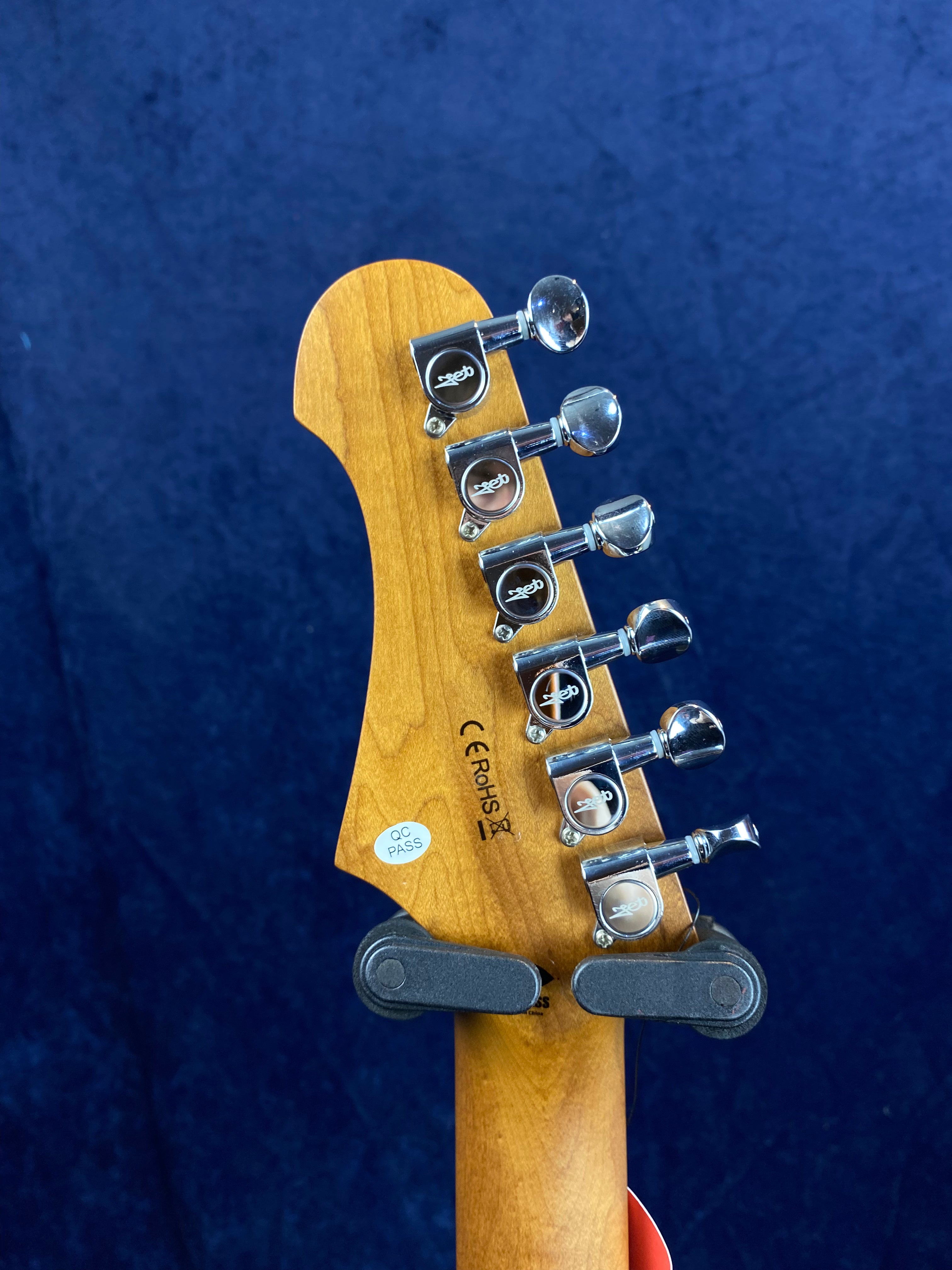 Six chrome tuning pegs on a light wood electric guitar headstock, with a QC Pass sticker and CE RoHS markings visible against a dark blue background.