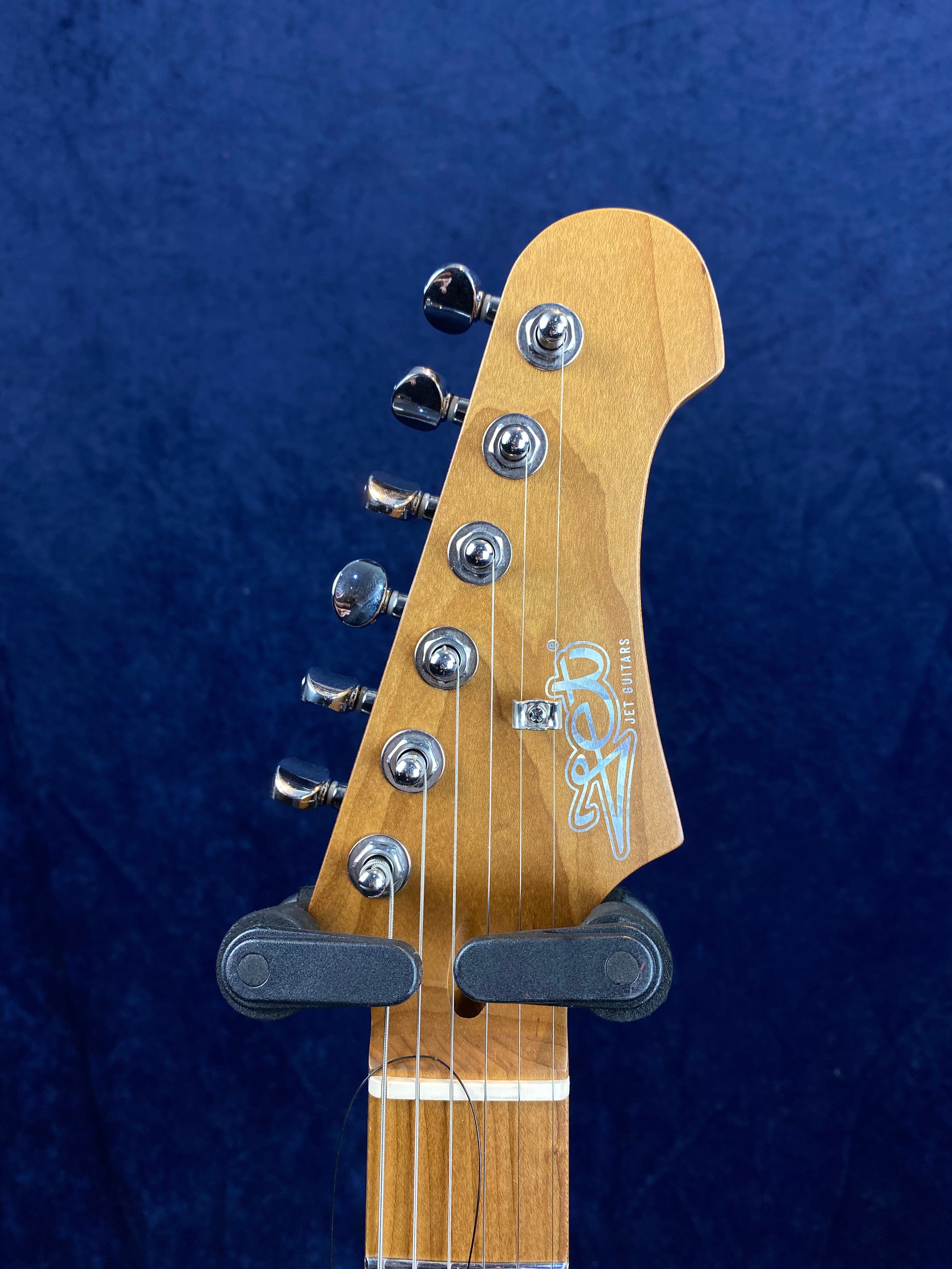 Light brown electric guitar headstock with six chrome tuning pegs, Jet Guitars logo, and strings, set against a dark blue background.