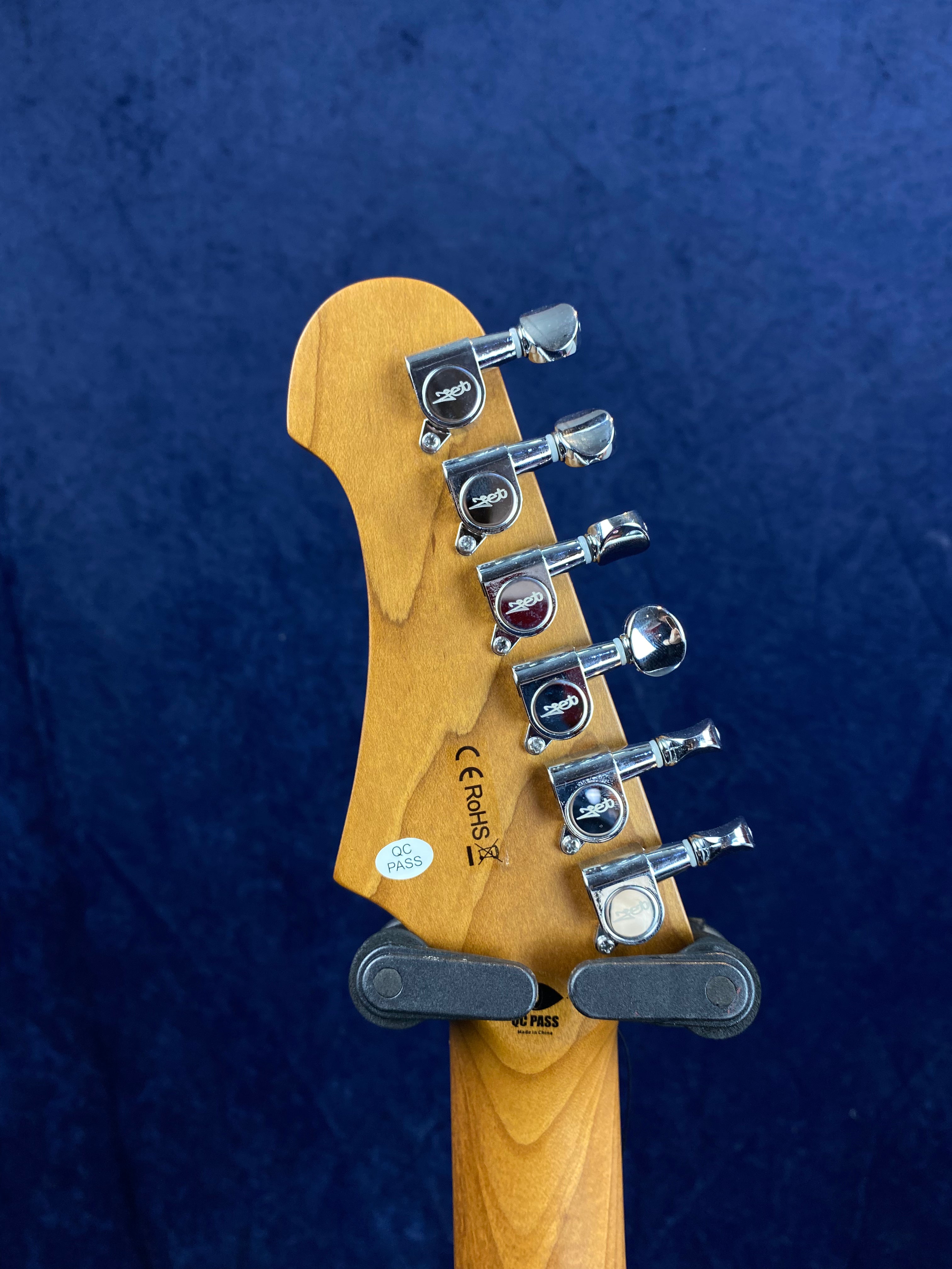Six chrome tuning pegs on a light wood electric guitar headstock, with QC and certification stickers, held by a guitar hanger against a dark blue background.