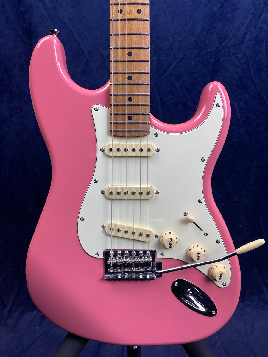 Pink electric guitar with a white pickguard, three cream single-coil pickups, a tremolo bridge, and maple neck with dot inlays.