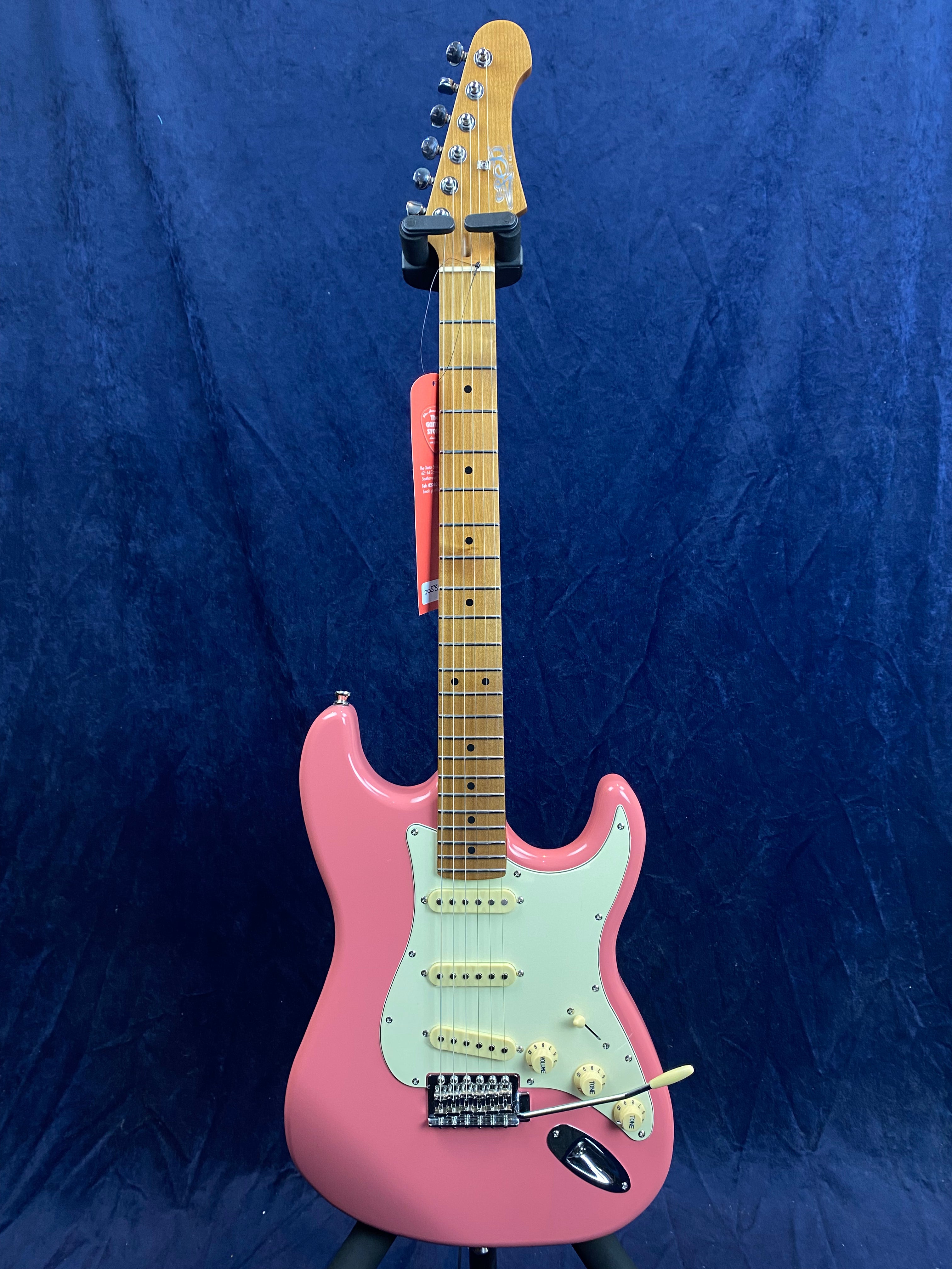 Pink electric guitar with a white pickguard, maple neck, three single-coil pickups, and a tremolo arm, displayed upright against a dark blue background.