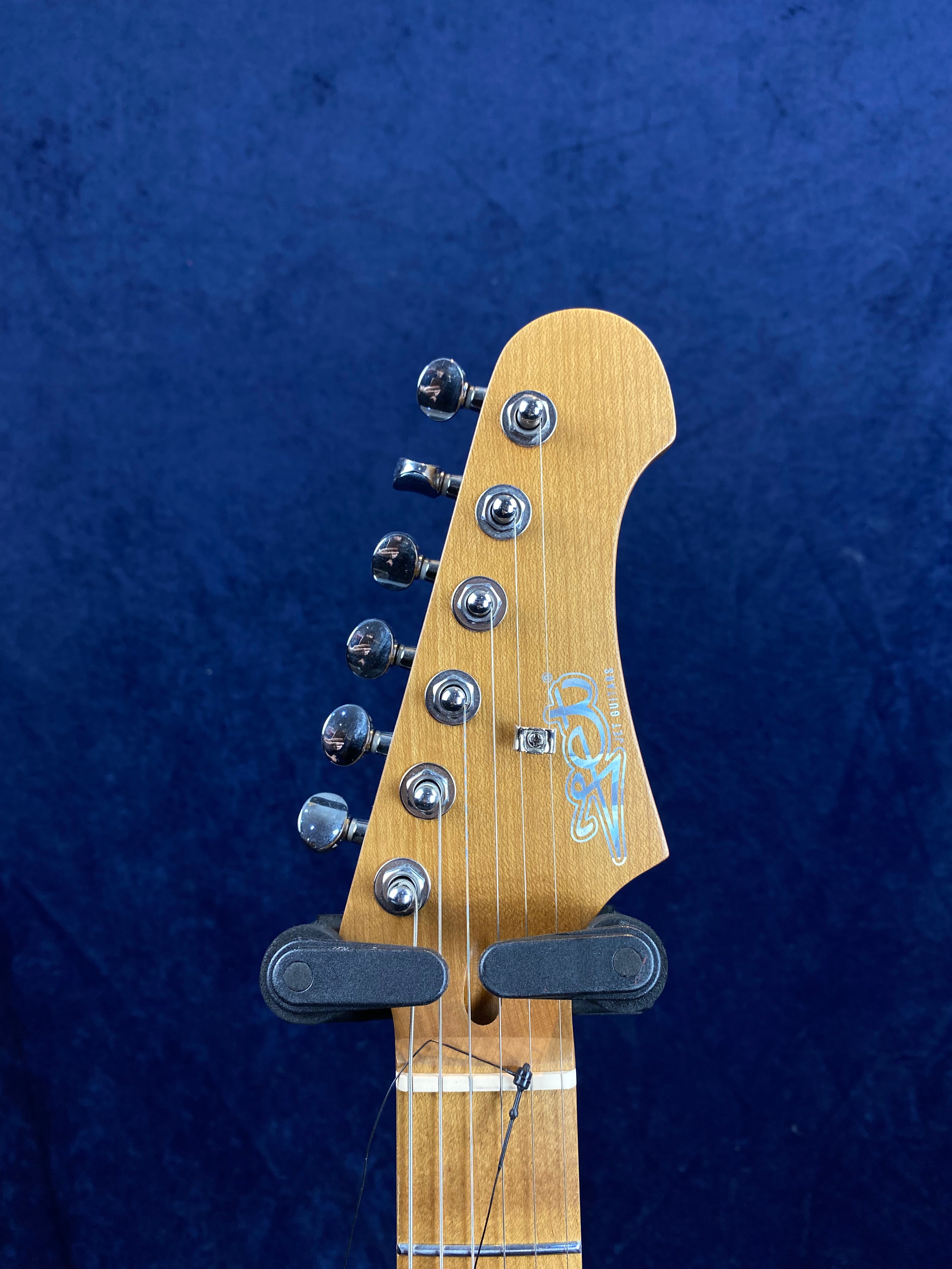 Six tuning pegs on a light wood electric guitar headstock with a logo, held upright in a black guitar hanger against a blue background.