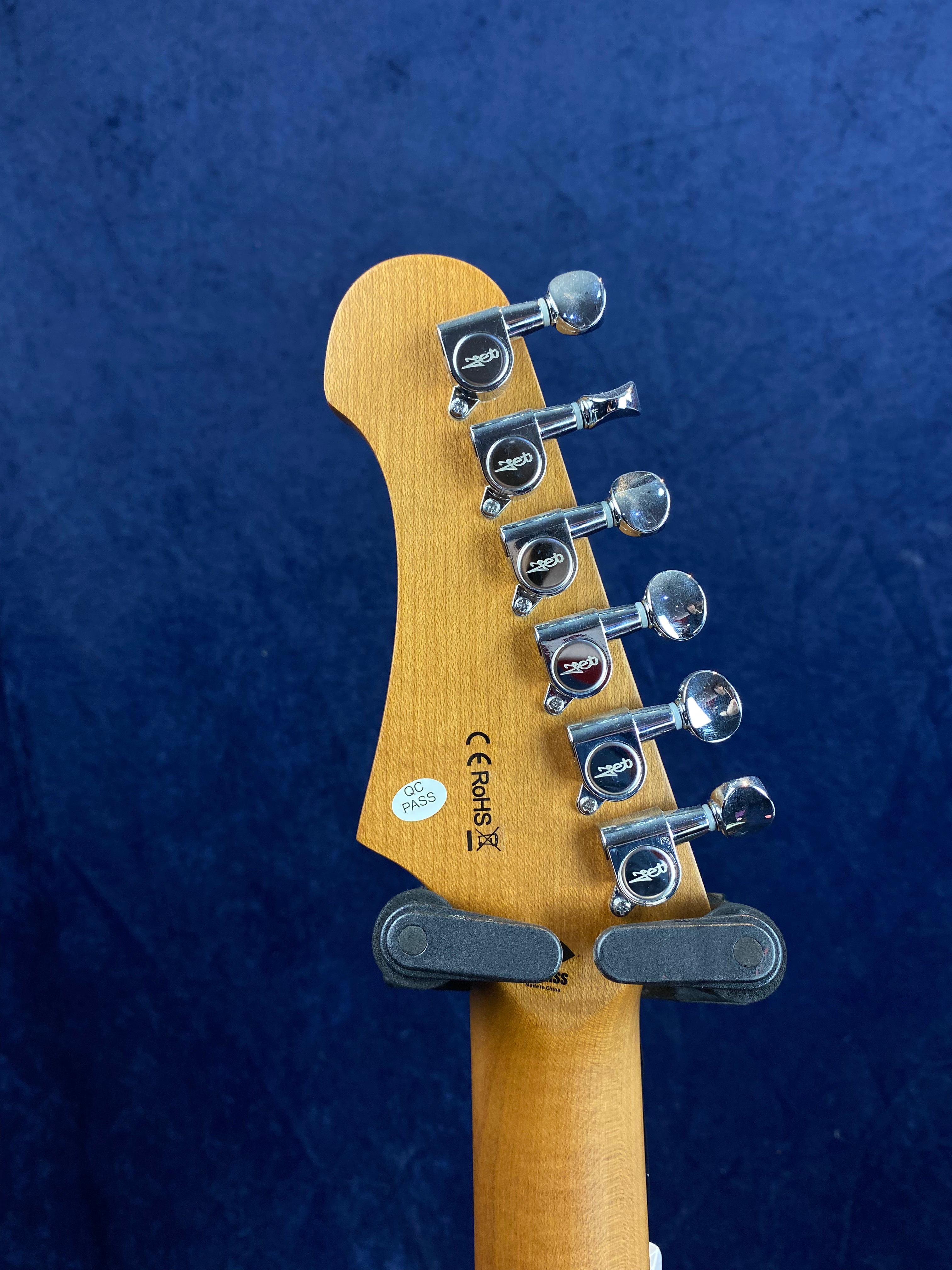 Six tuning pegs on a wooden electric guitar headstock, with certification stickers and a guitar hanger gripping the neck, blue background.