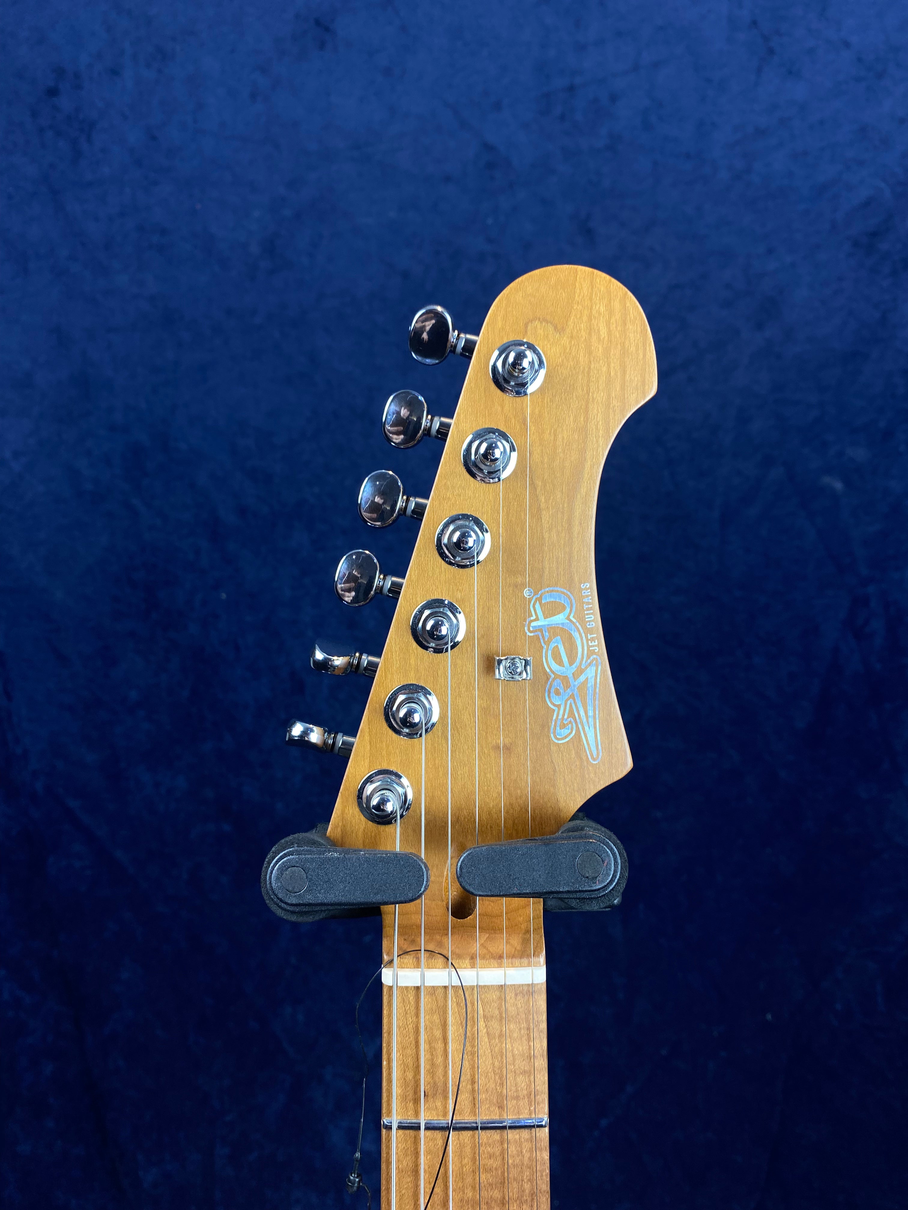 Natural wood electric guitar headstock with six chrome tuning pegs, a string tree, and blue logo, set against a dark blue background.