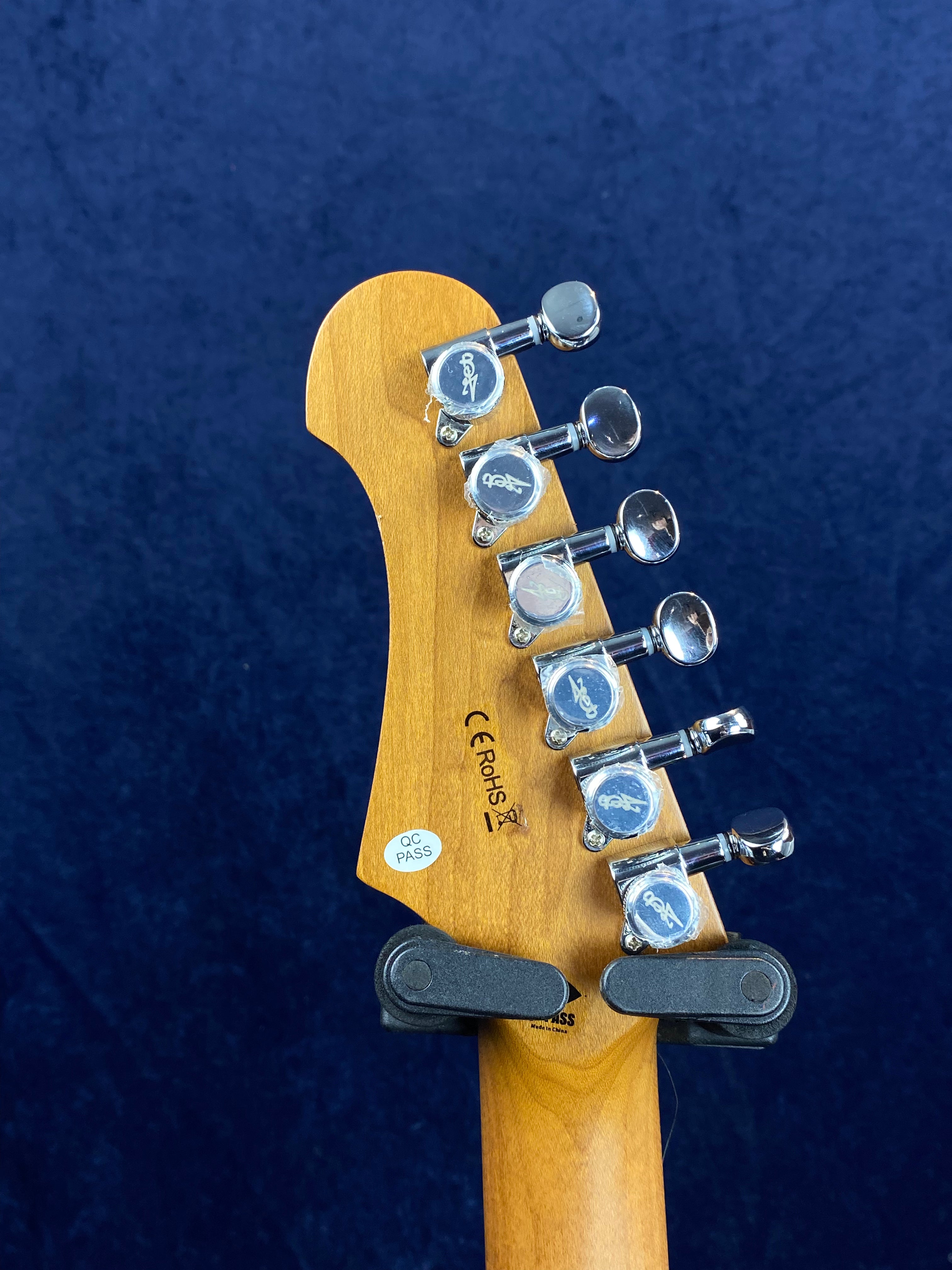 Six chrome tuning pegs on a natural wood electric guitar headstock, with QC PASS sticker and CE RoHS markings visible against a dark blue background.