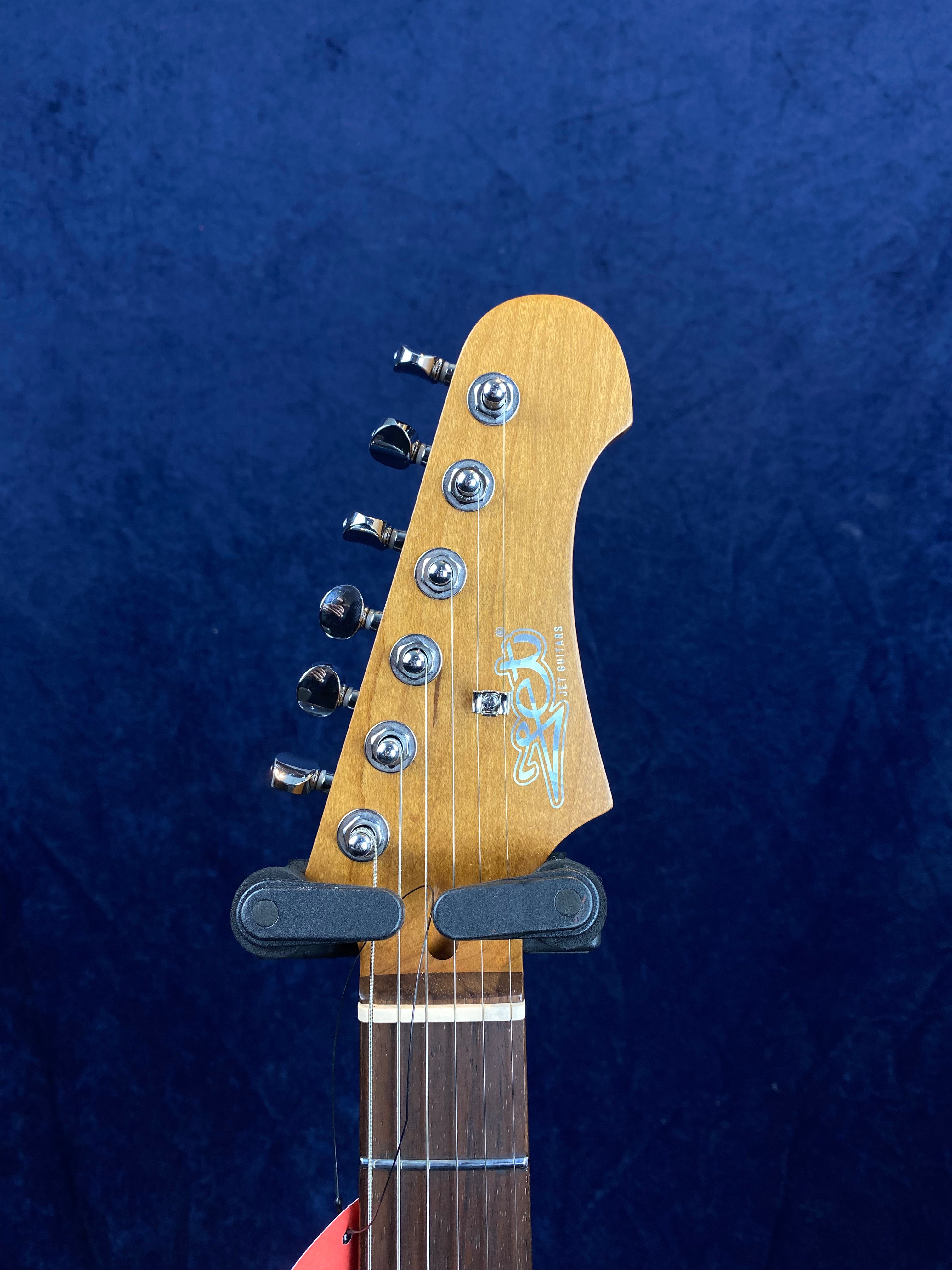 Light wood electric guitar headstock with six chrome tuning pegs, Jet Guitars logo, and dark wood fretboard, against a dark blue background.