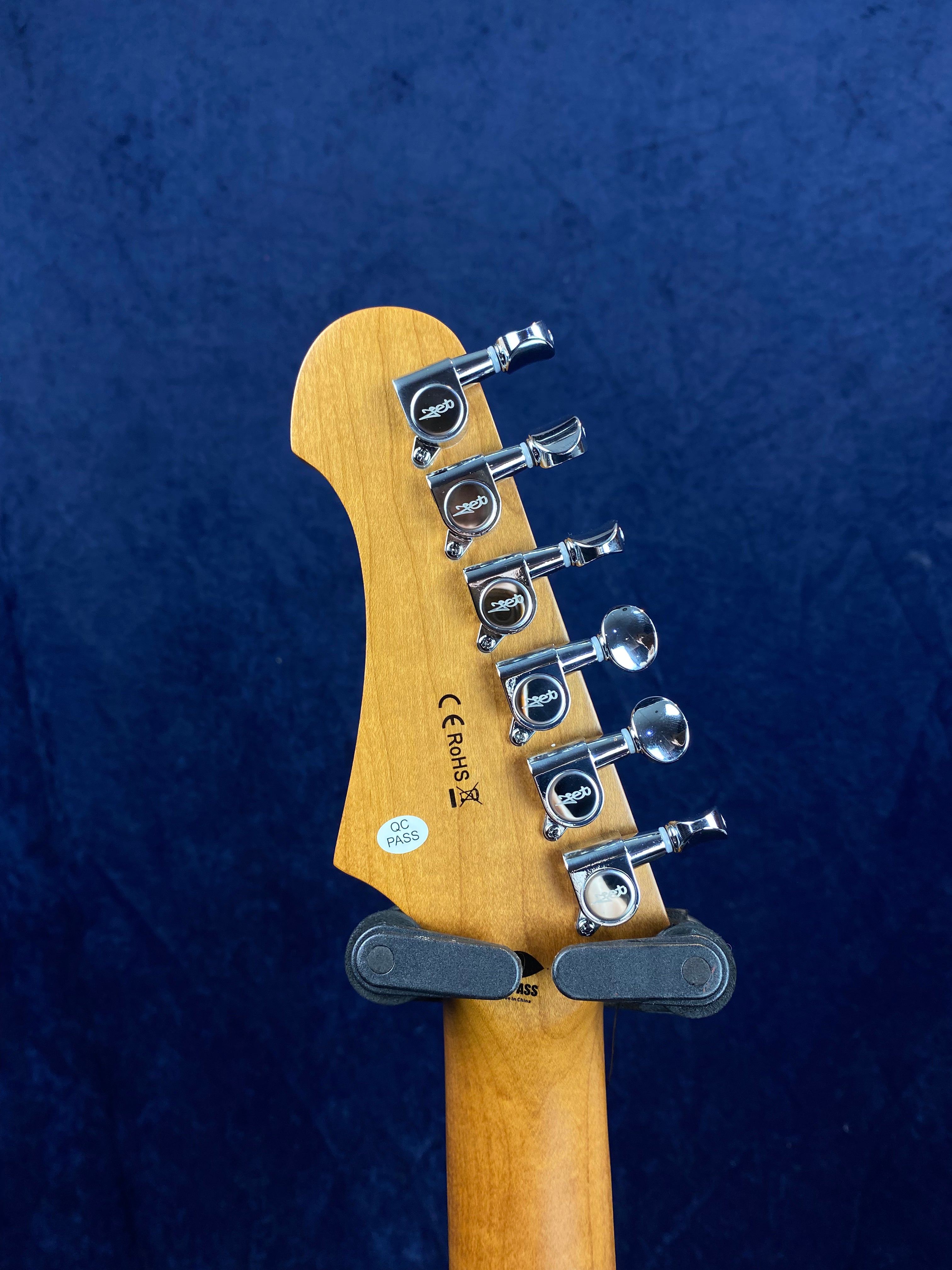 Electric guitar headstock with six chrome tuning pegs, “QC PASS” sticker, and wood grain visible against a dark blue background.