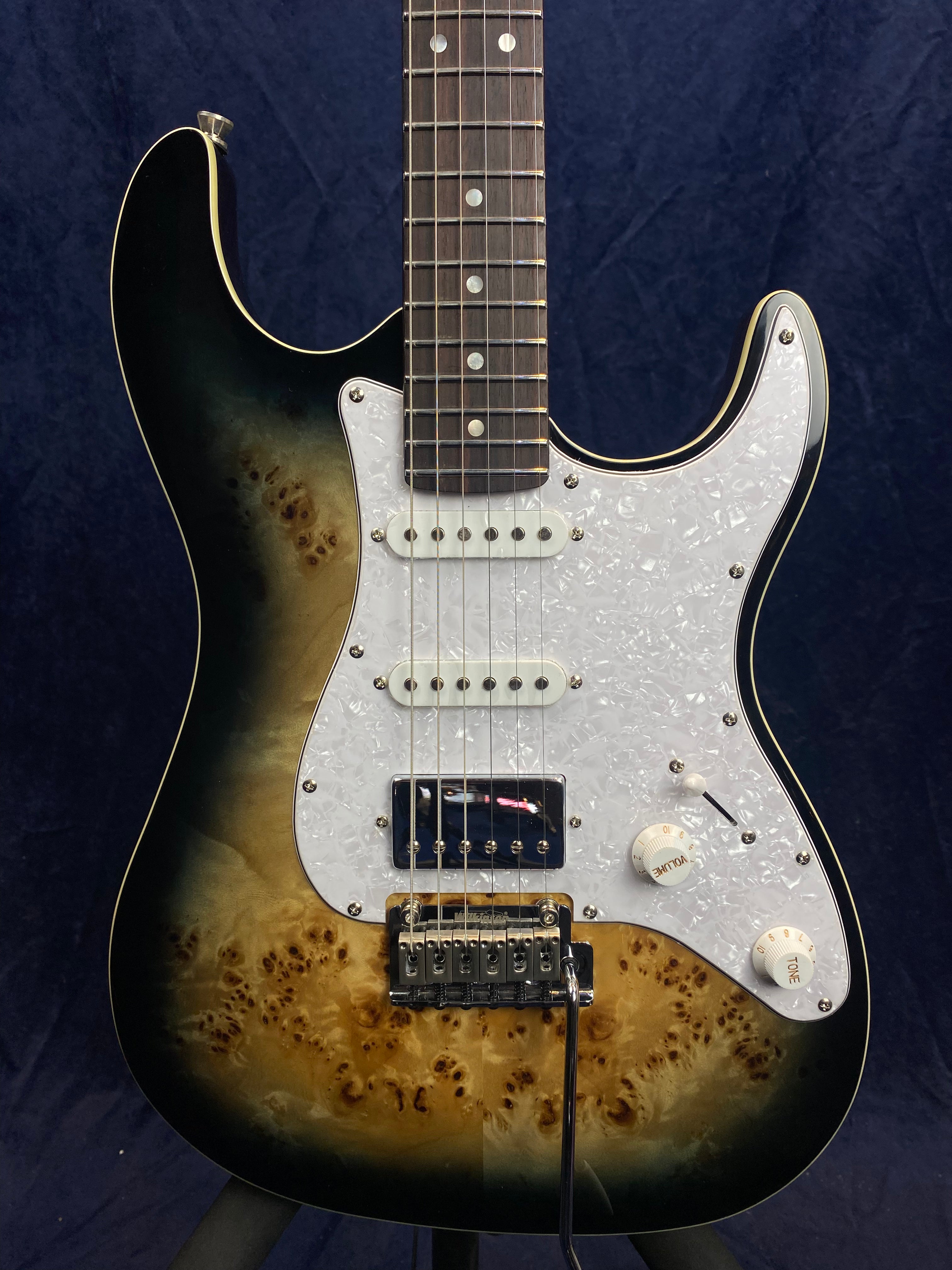 Electric guitar with a glossy sunburst and burled wood finish, white pearl pickguard, two single-coil pickups, one humbucker, and whammy bar.