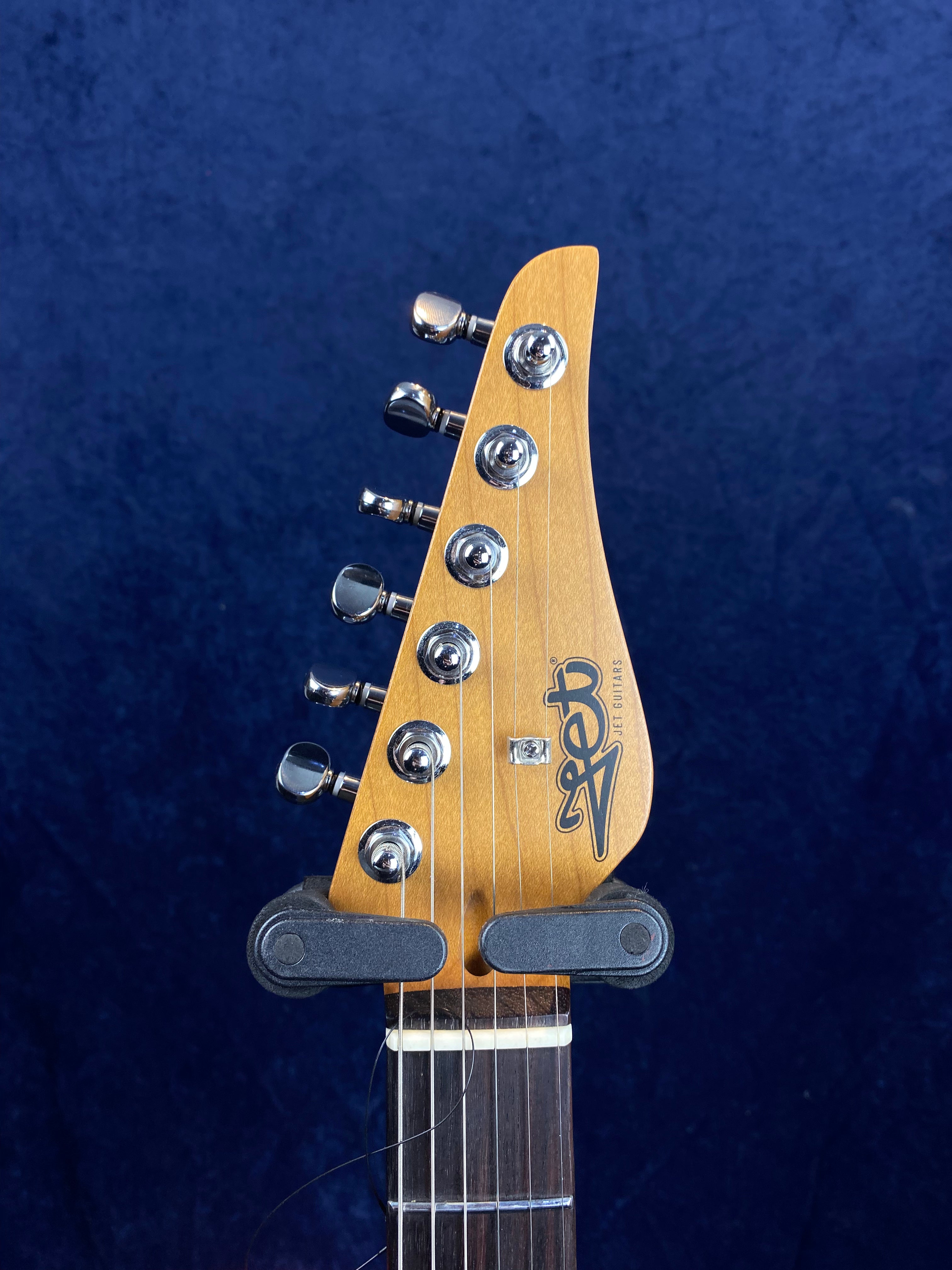 Six chrome tuning pegs on a light wood Jet Guitars electric guitar headstock, strings attached, dark fretboard below, blue background.