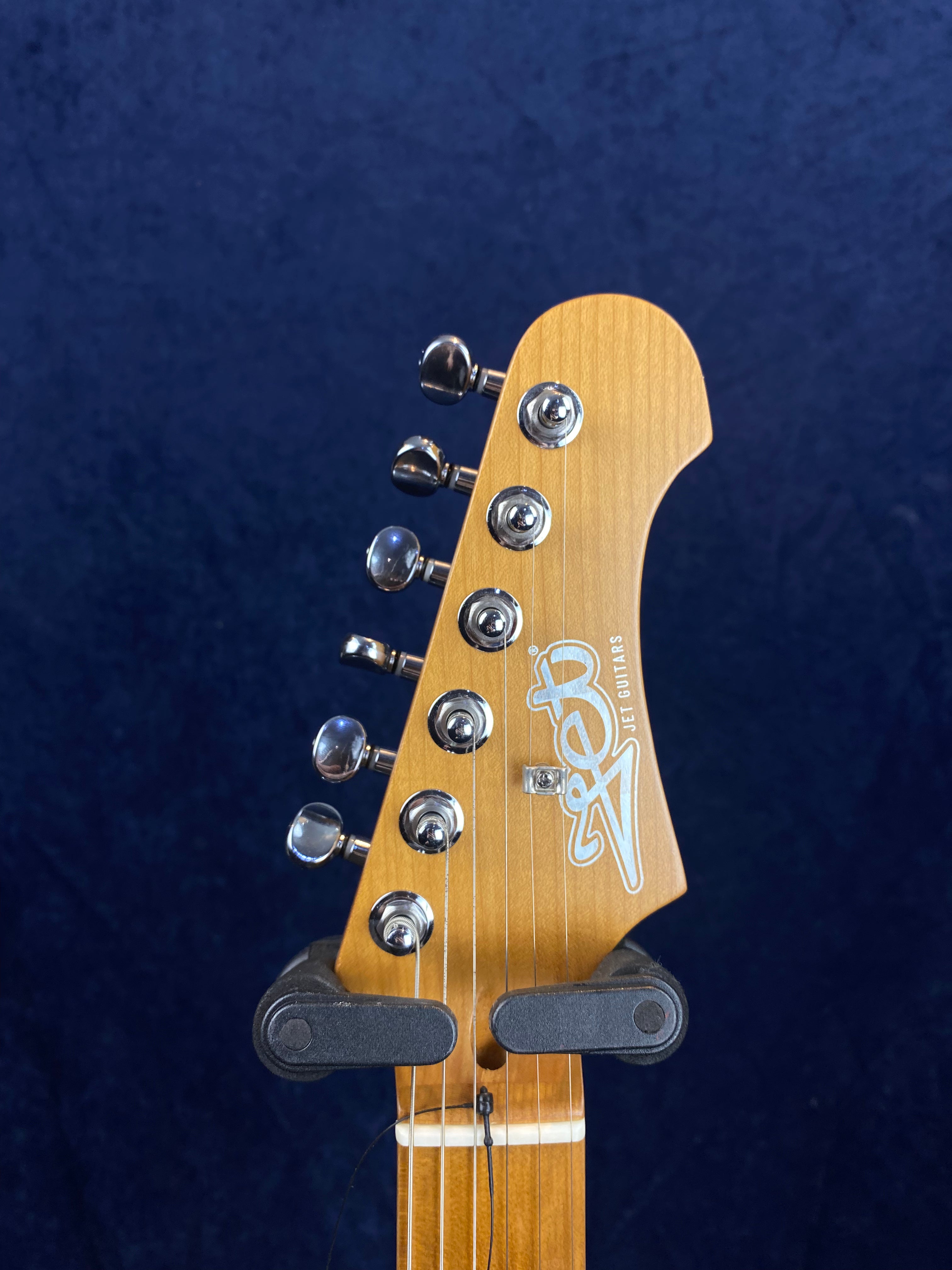 Light wood electric guitar headstock with six chrome tuning pegs and “Jet Guitars” logo, mounted on a black guitar holder.