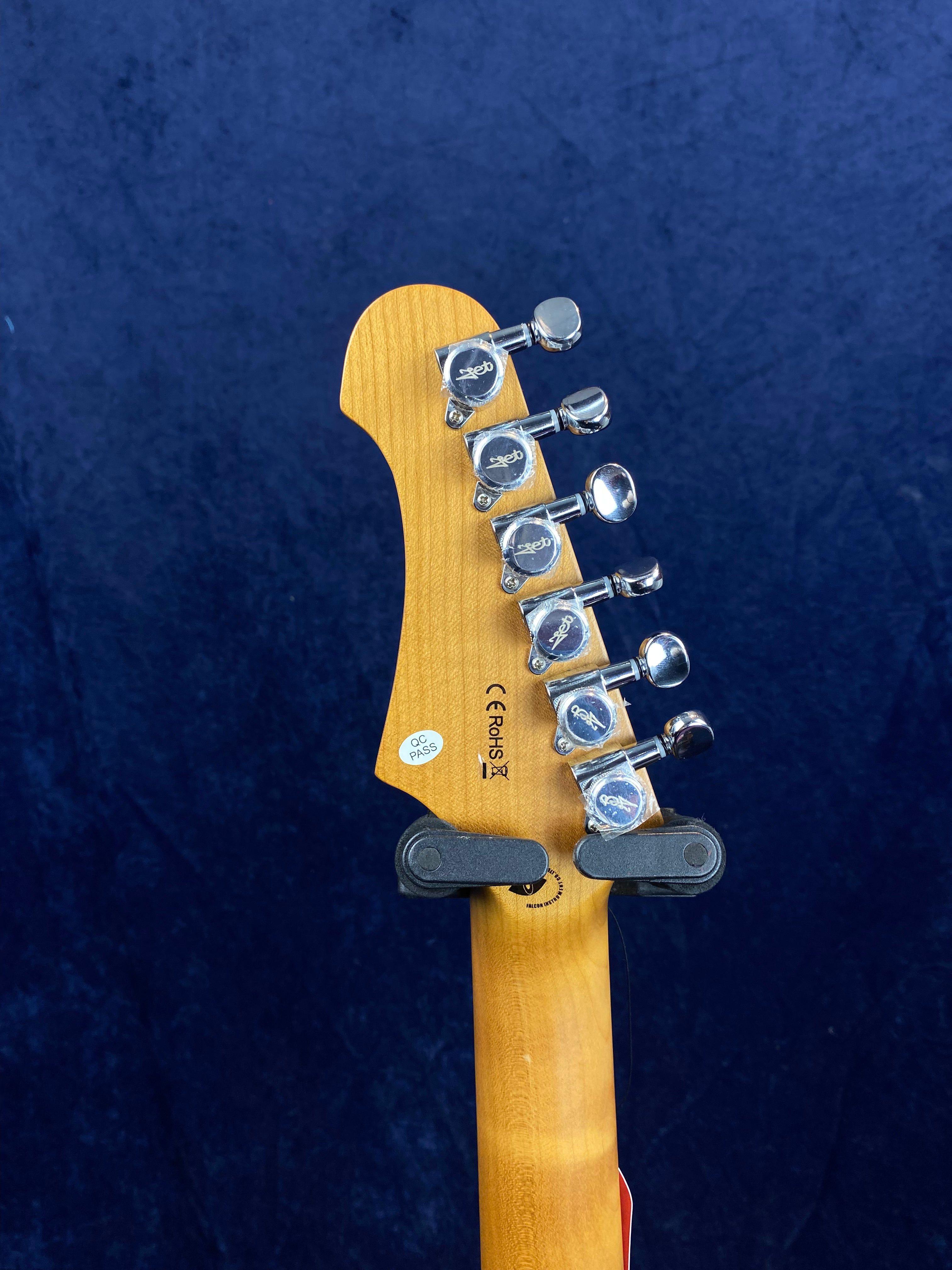 Electric guitar headstock in natural wood with six silver tuning pegs, certification stickers, and mounted on a black hanger against a dark blue background.