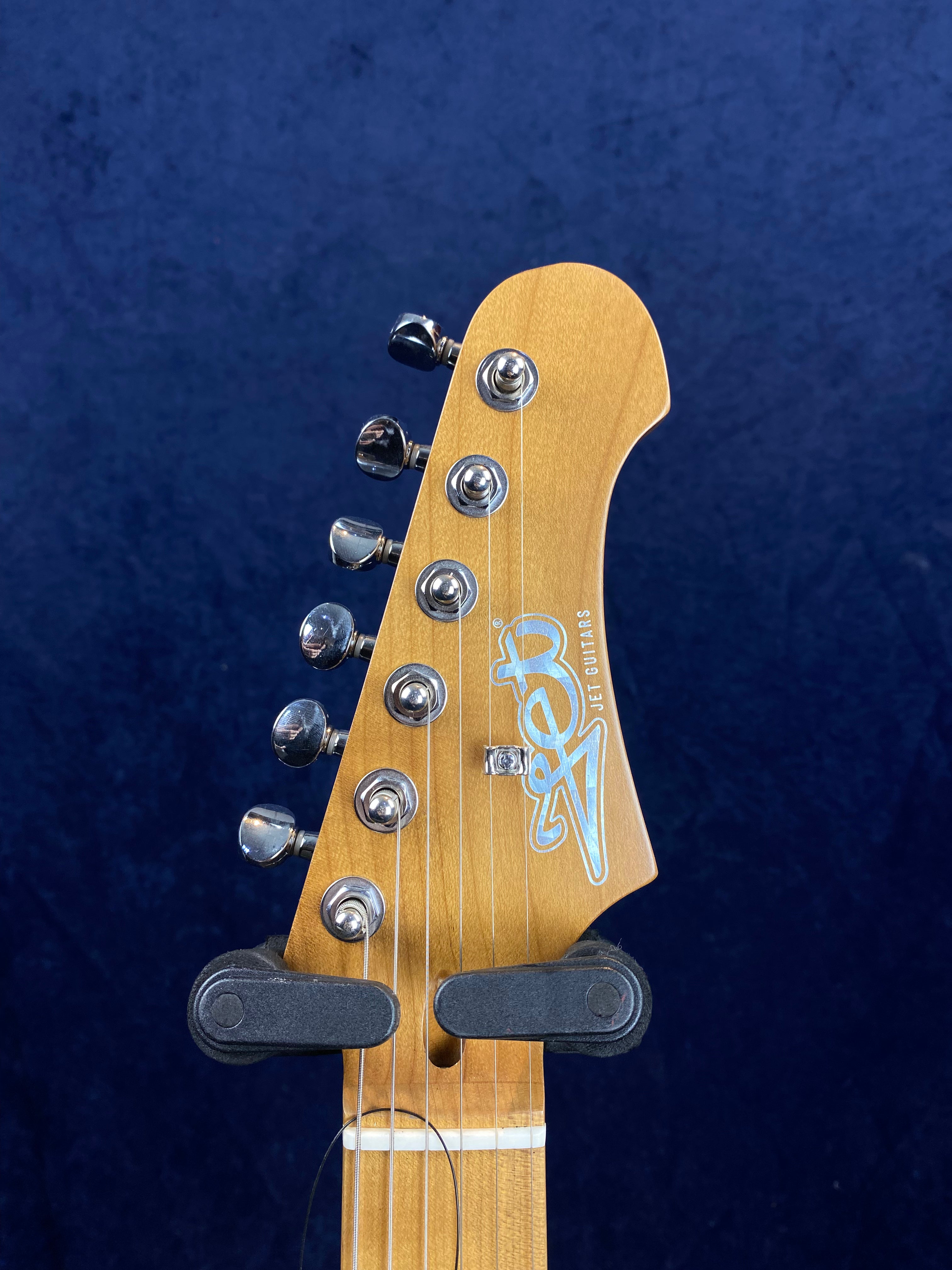 Light wood electric guitar headstock with six chrome tuning pegs and "Jet Guitars" logo on a dark blue background.