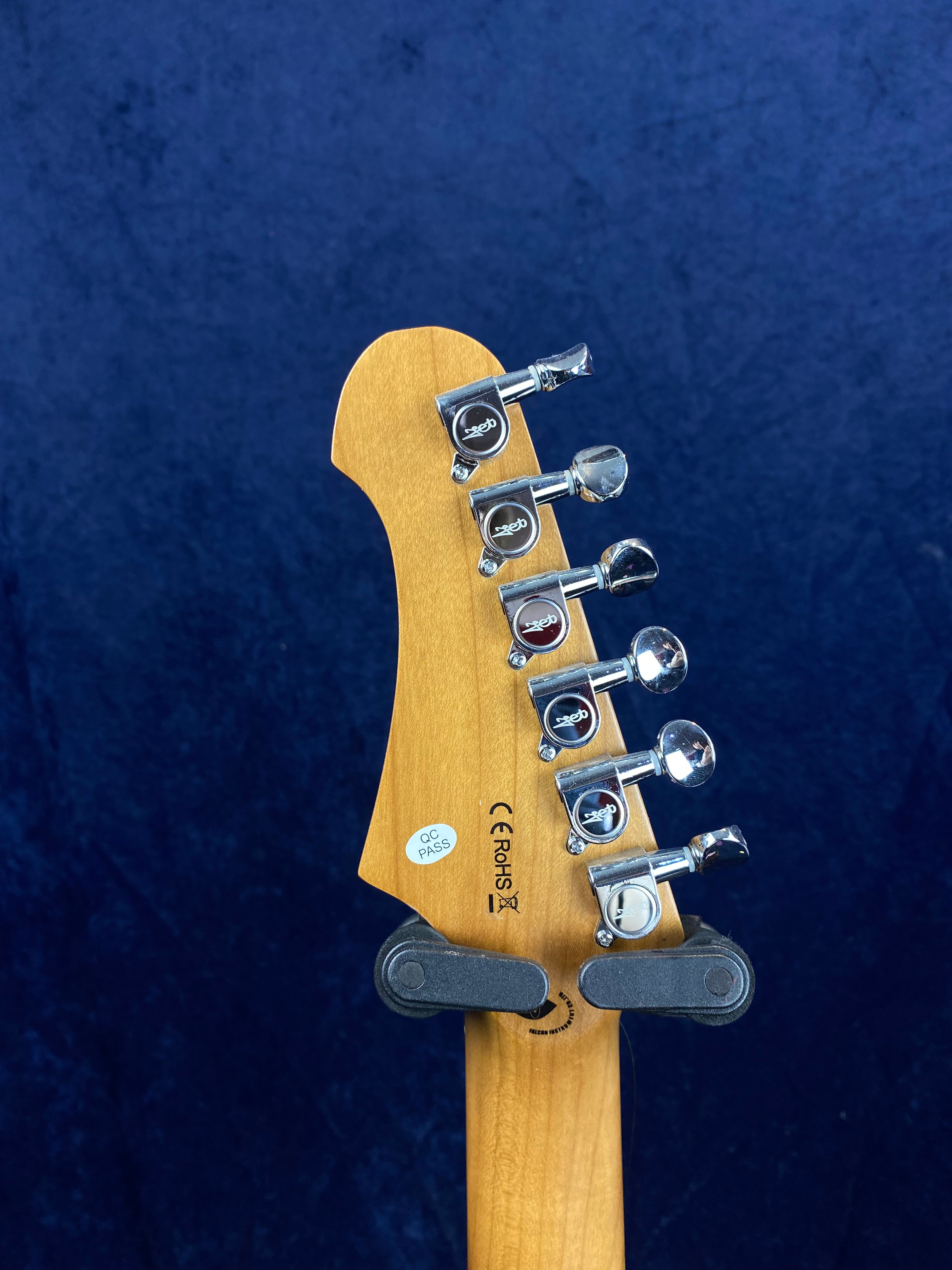 Six chrome tuning pegs on the back of a light wood electric guitar headstock, with certification stickers and a black padded wall mount.