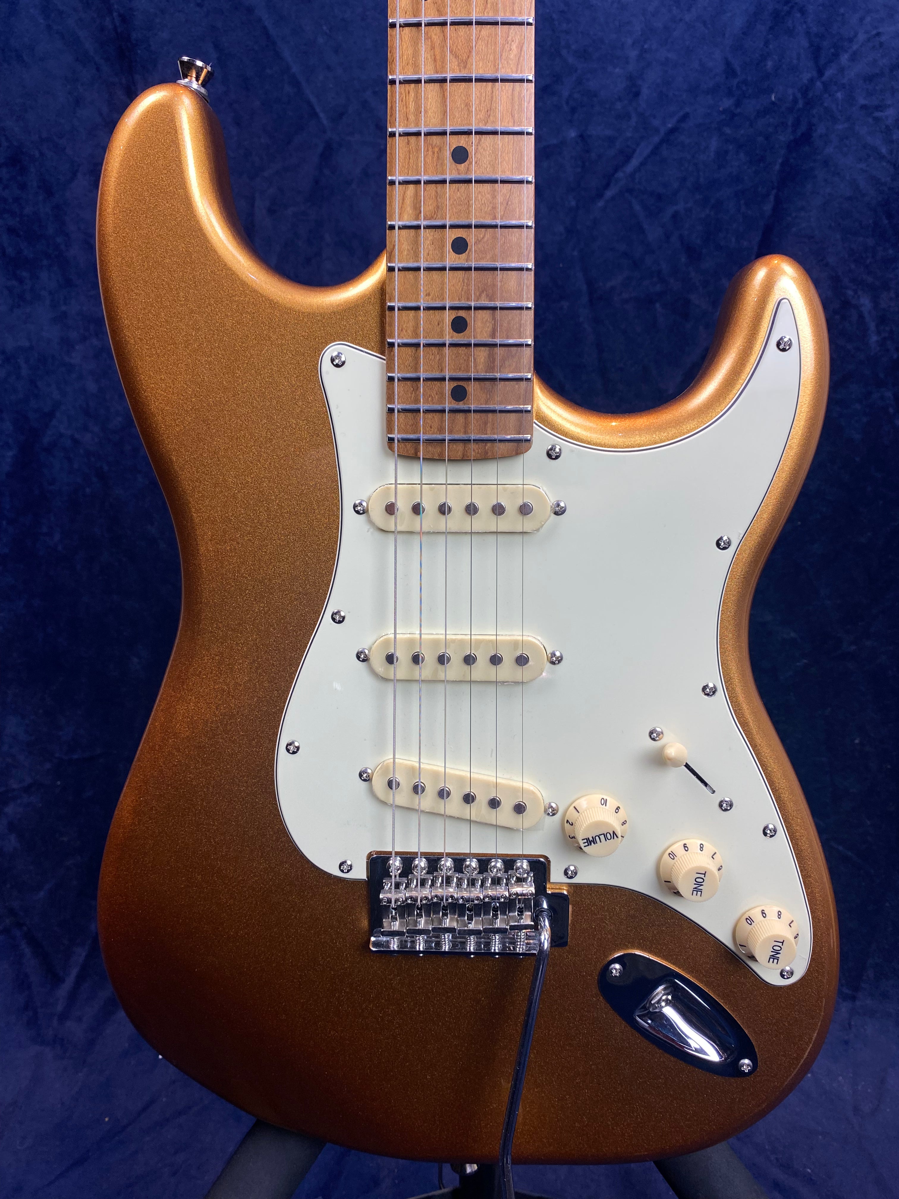 Metallic bronze electric guitar with white pickguard, three single-coil pickups, three control knobs, and a wooden neck against a dark background.