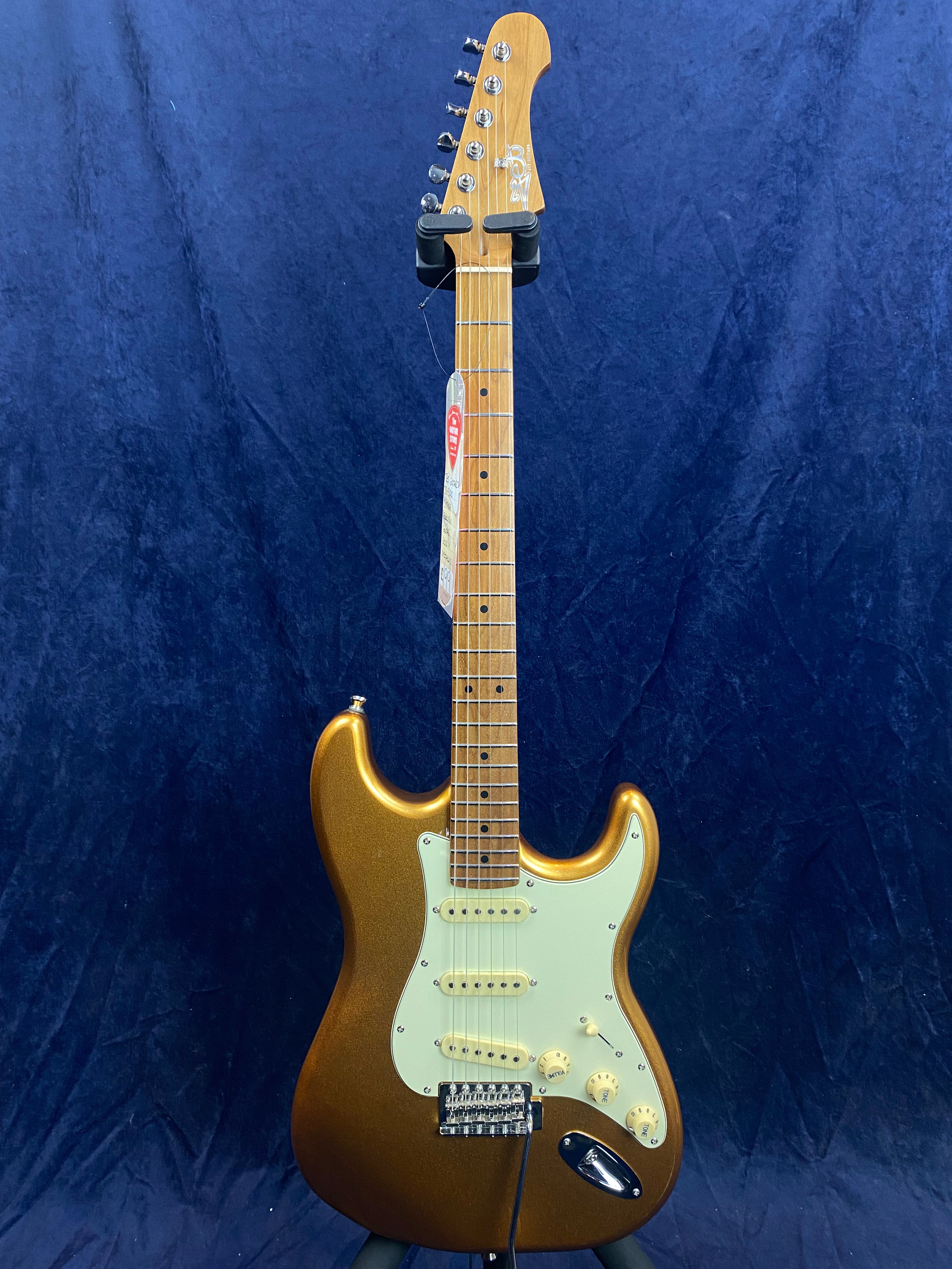 Metallic gold electric guitar with a maple neck, white pickguard, three single-coil pickups, and control knobs, displayed against a dark blue background.