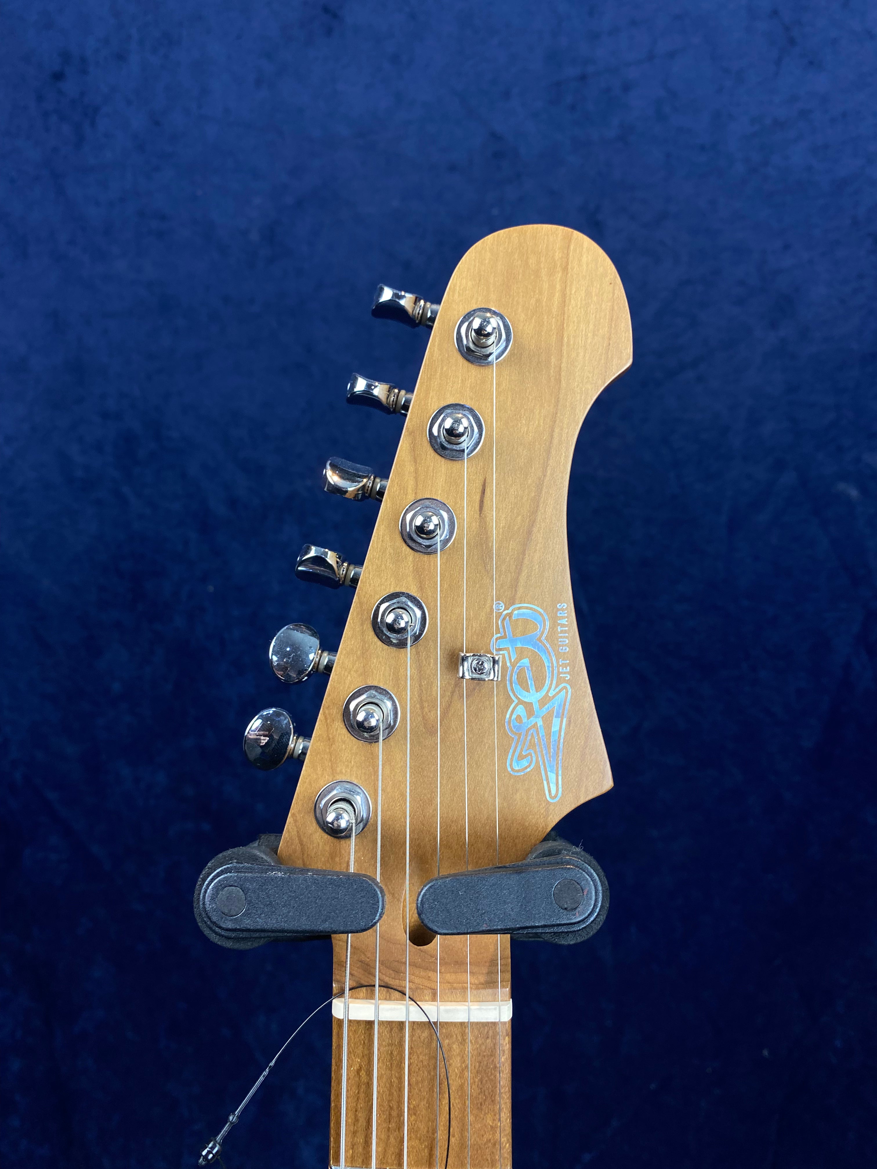 Light brown electric guitar headstock with six metal tuning pegs, blue Jet Guitars logo, and strings in a black wall mount, set against a dark blue background.