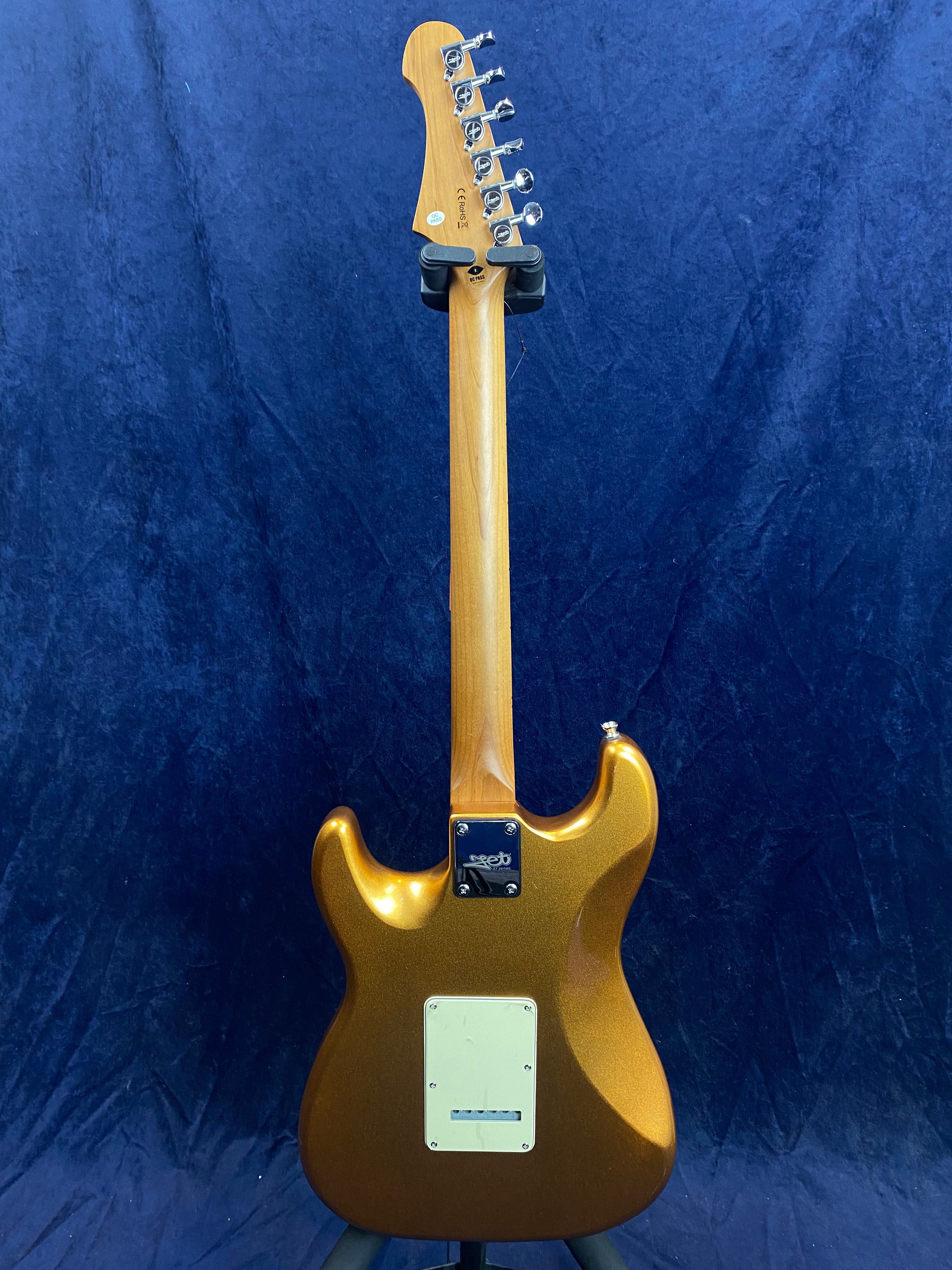 Electric guitar shown from the back with a metallic bronze body, light wood neck, chrome tuners, and a rectangular white plate on the lower body.
