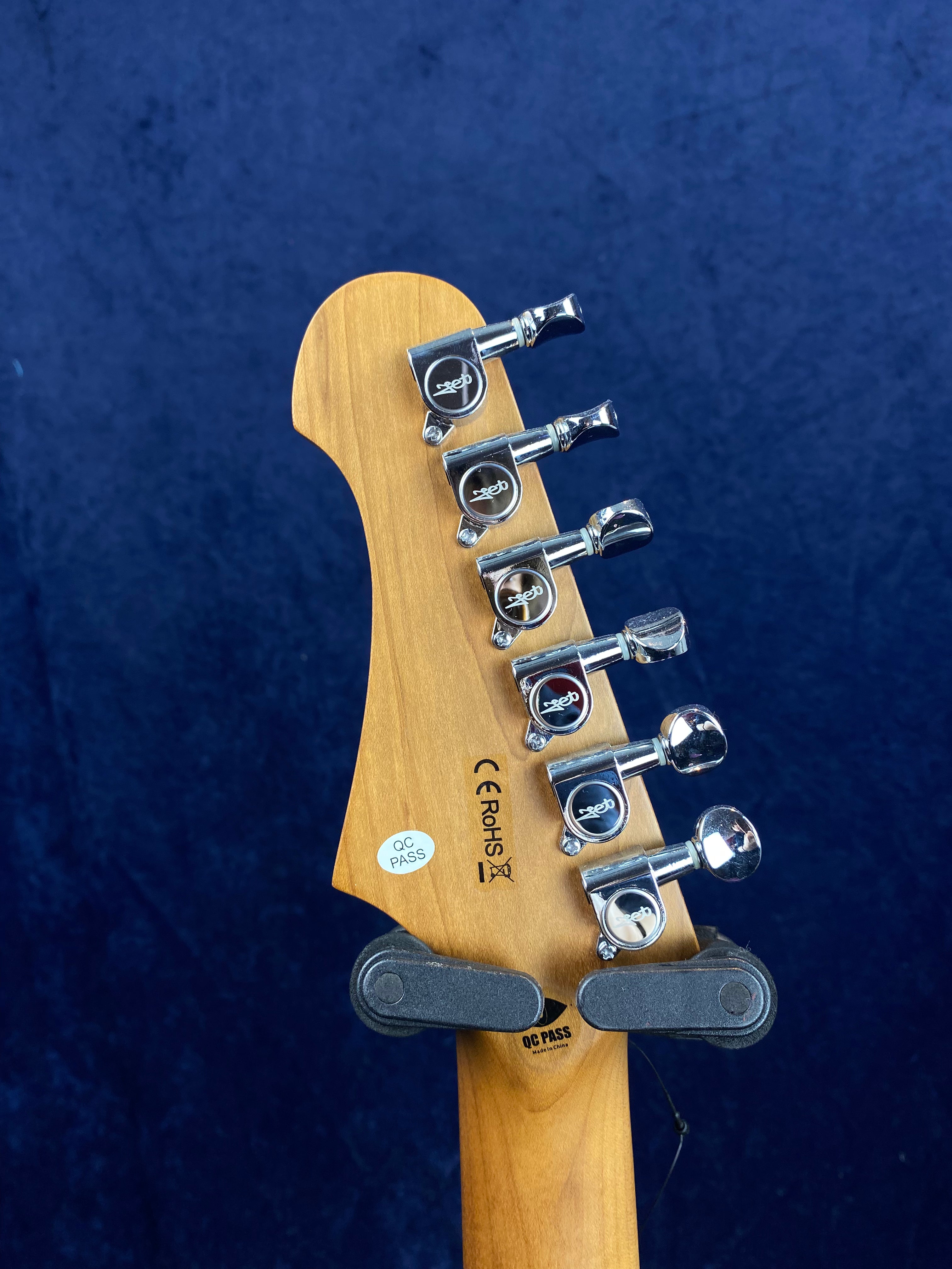 Six chrome tuning pegs on the back of a wooden electric guitar headstock, with QC and CE/RoHS certification stickers visible.