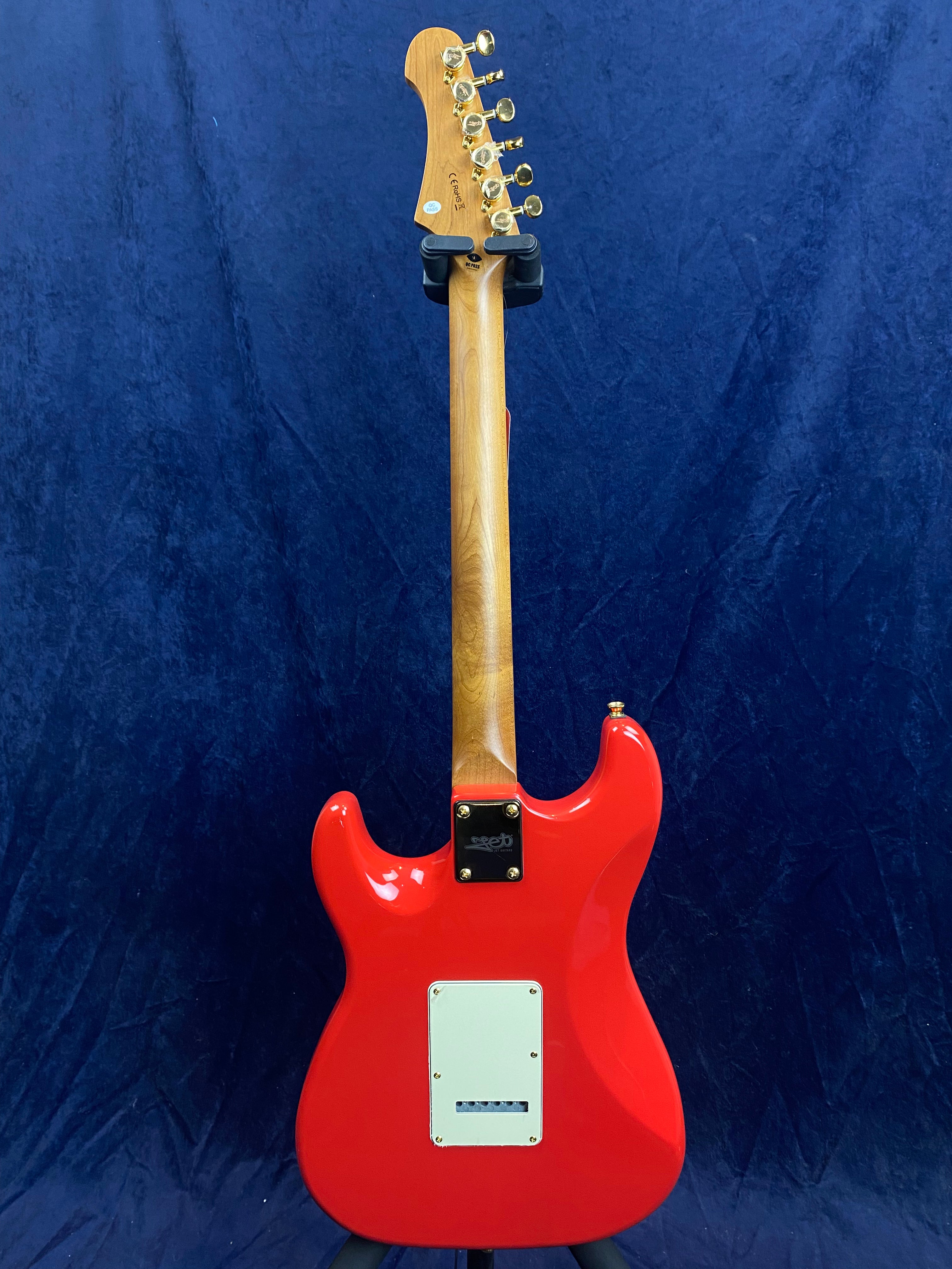 Bright red Jet electric guitar shown from the back, natural wood neck, gold tuning pegs, white backplate, set against a dark blue background.
