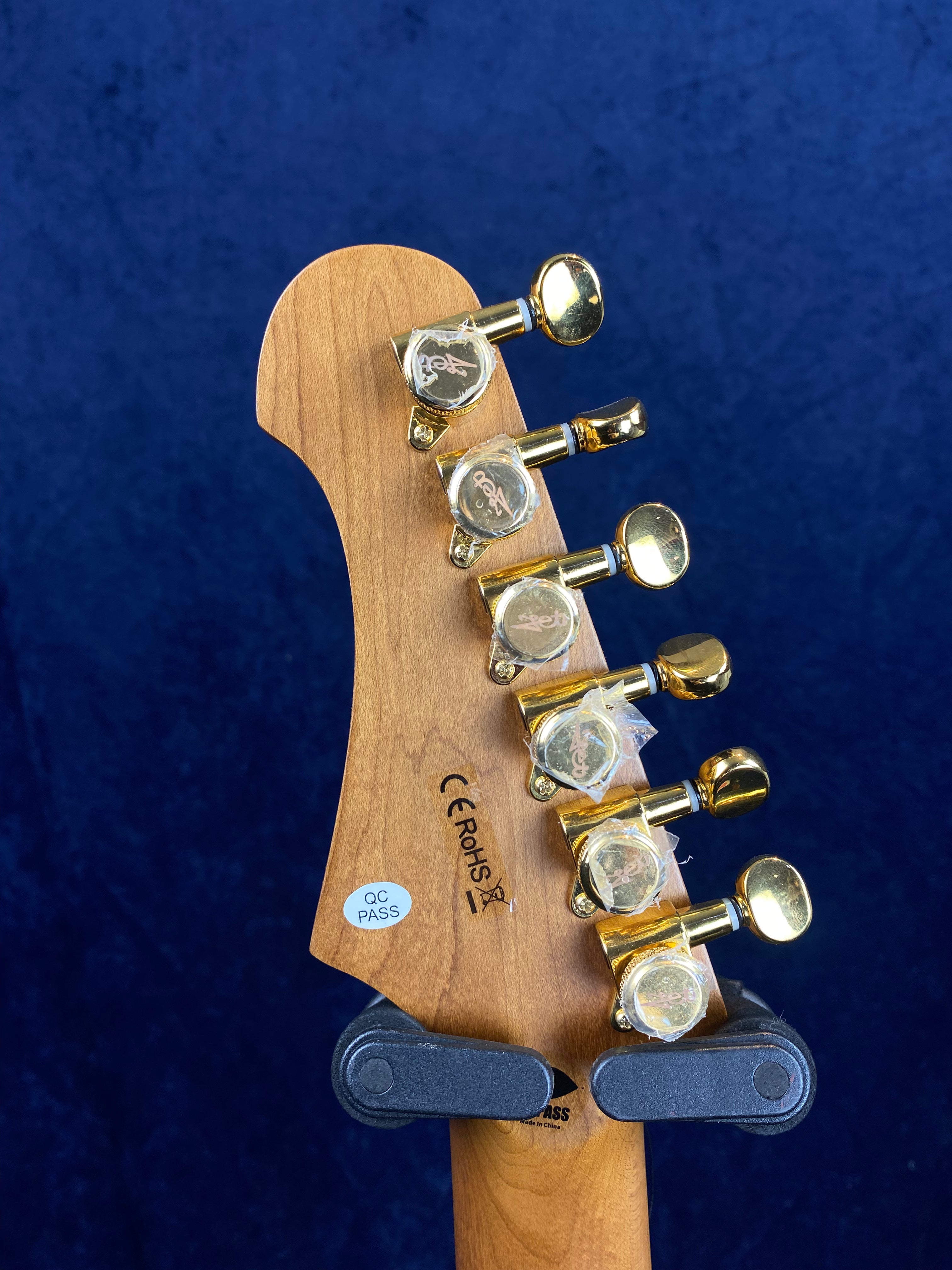 Jet Guitars headstock back with six gold tuning pegs, QC PASS sticker, and certification marks against a dark blue background.