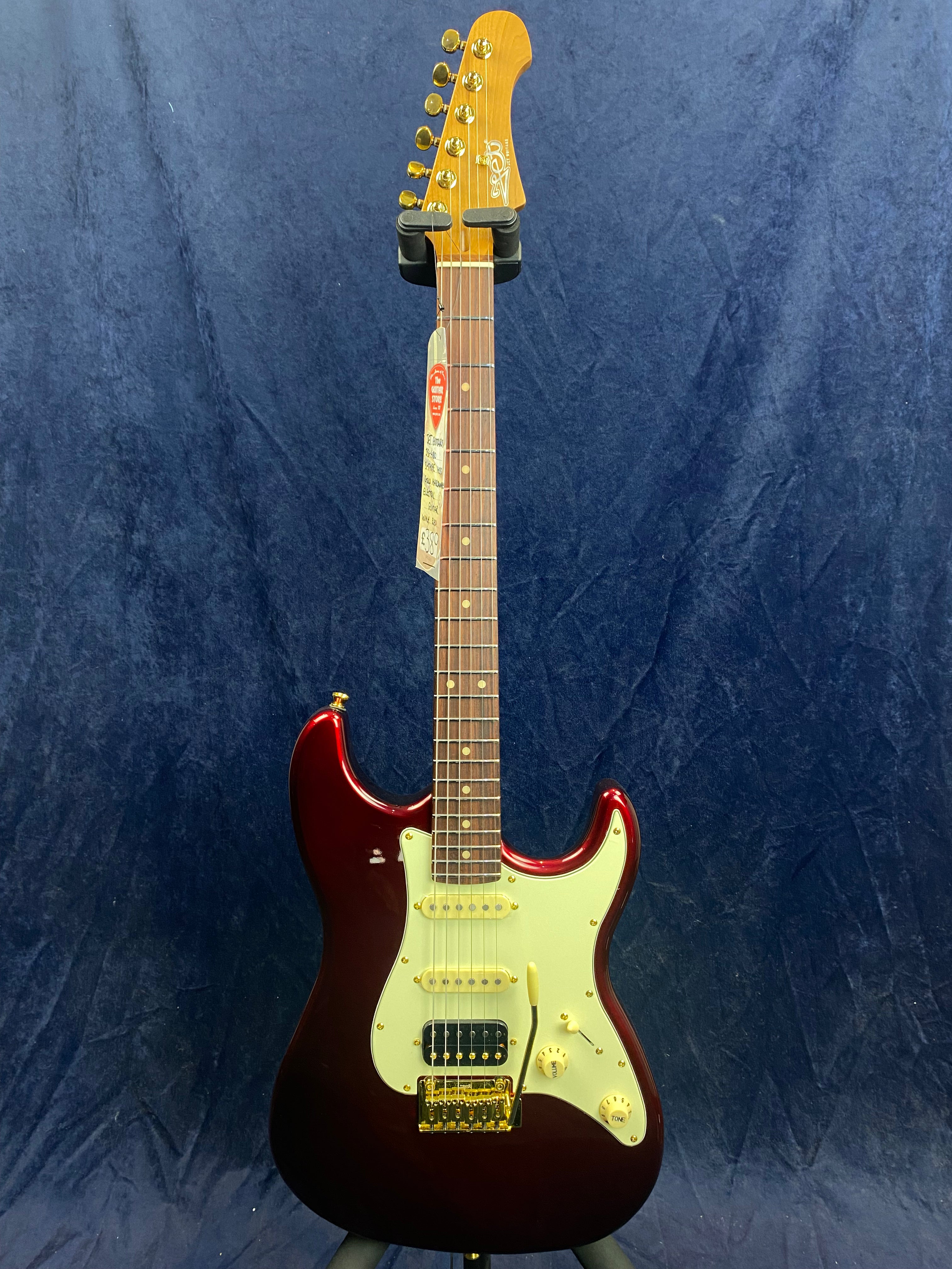 Red electric guitar with a white pickguard, gold hardware, and six tuning pegs, displayed upright against a dark blue background.