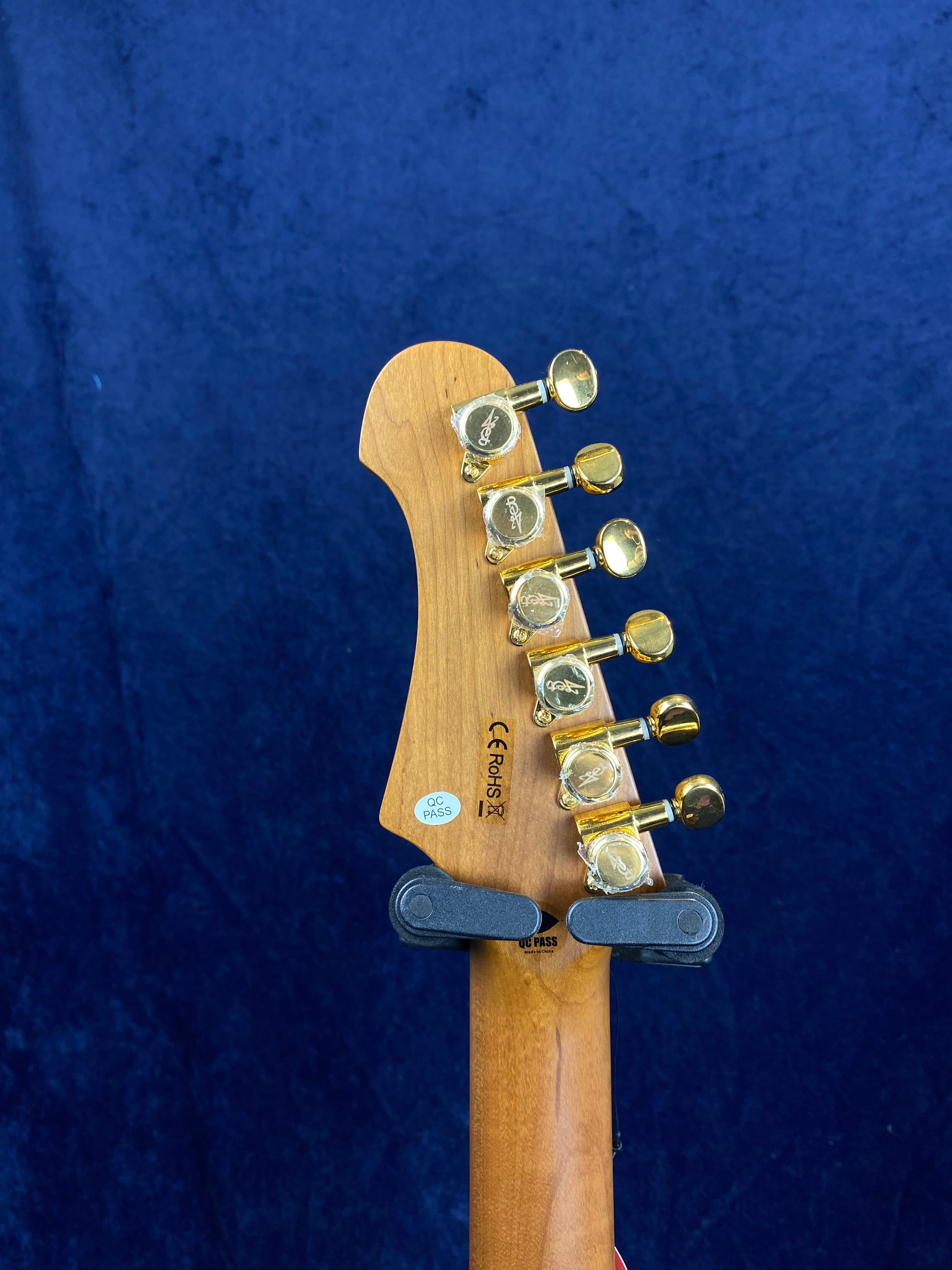 Electric guitar headstock with six gold tuning pegs, wooden finish, and quality control stickers, shown against a dark blue background.
