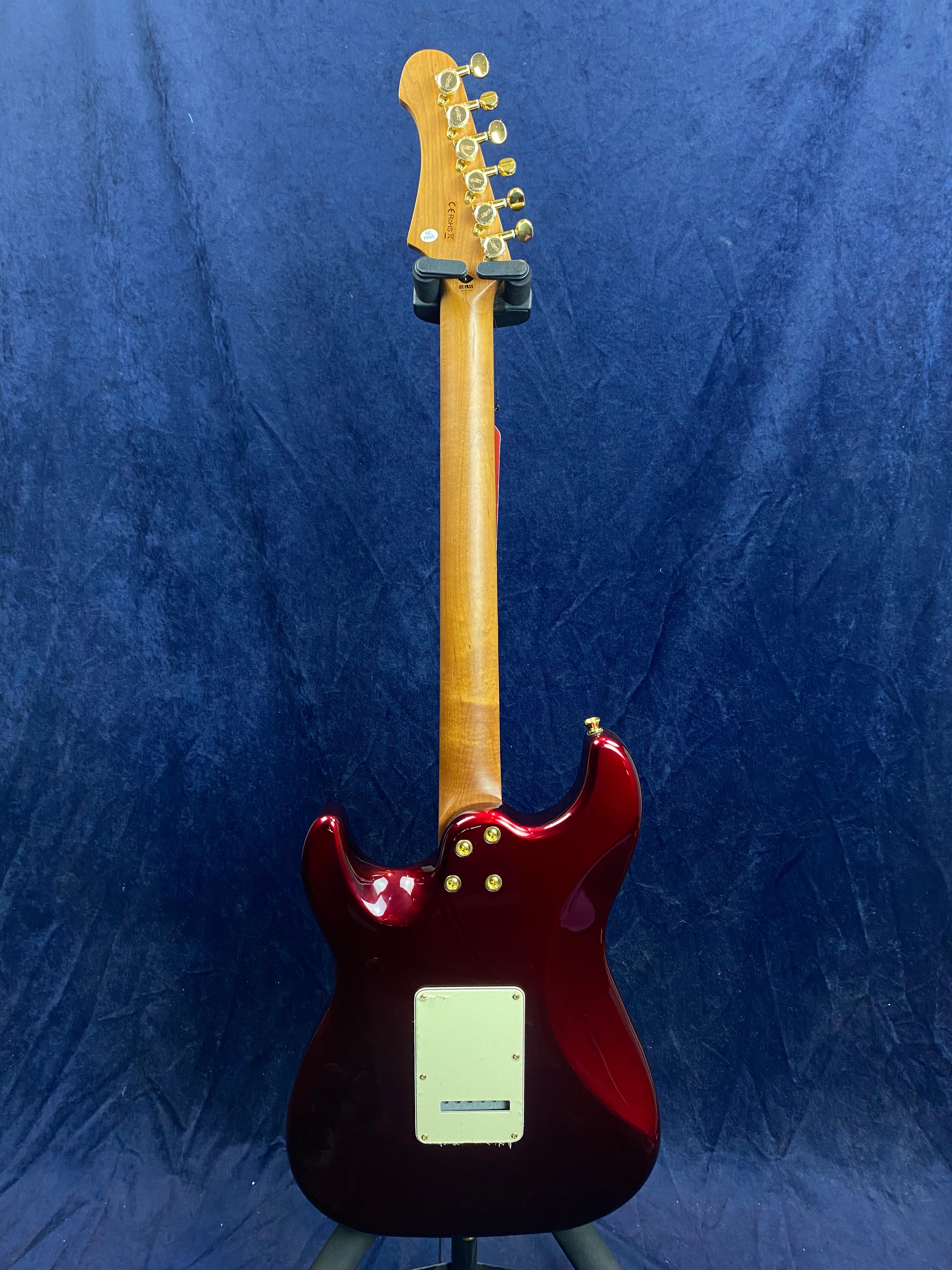 Dark red electric guitar shown from the back, with a light wood neck, gold tuning pegs, and a rectangular cream plate near the base.