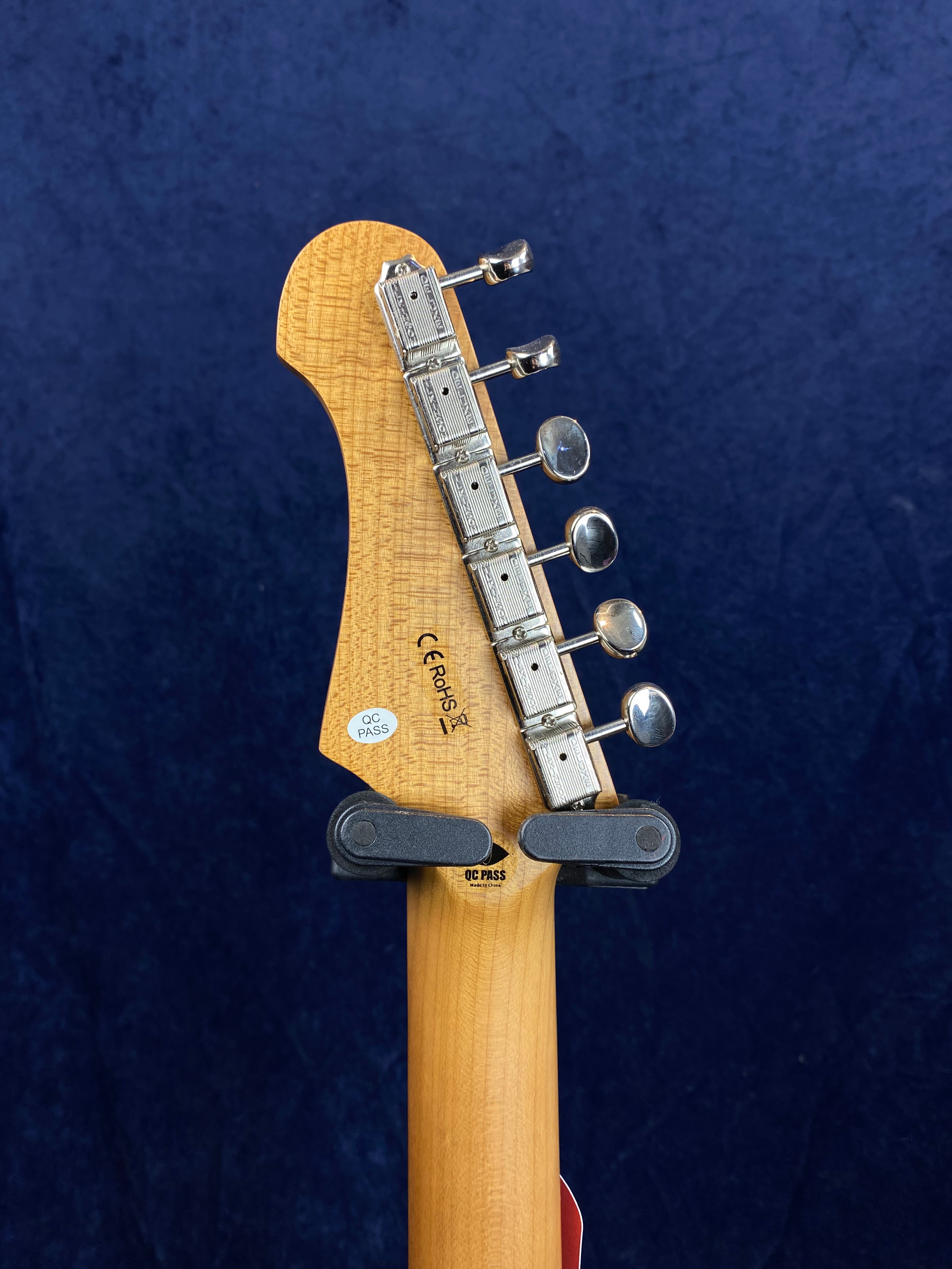 Electric guitar headstock with six chrome tuning pegs, a light wood finish, and quality control stickers against a dark blue background.