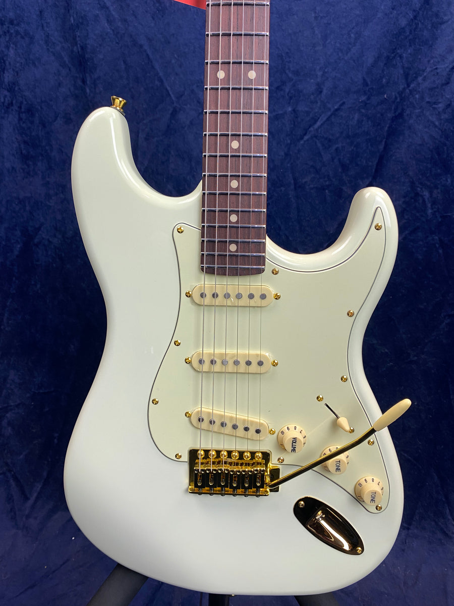 White electric guitar with three single-coil pickups, a rosewood fretboard, gold hardware, and cream control knobs against a dark blue background.
