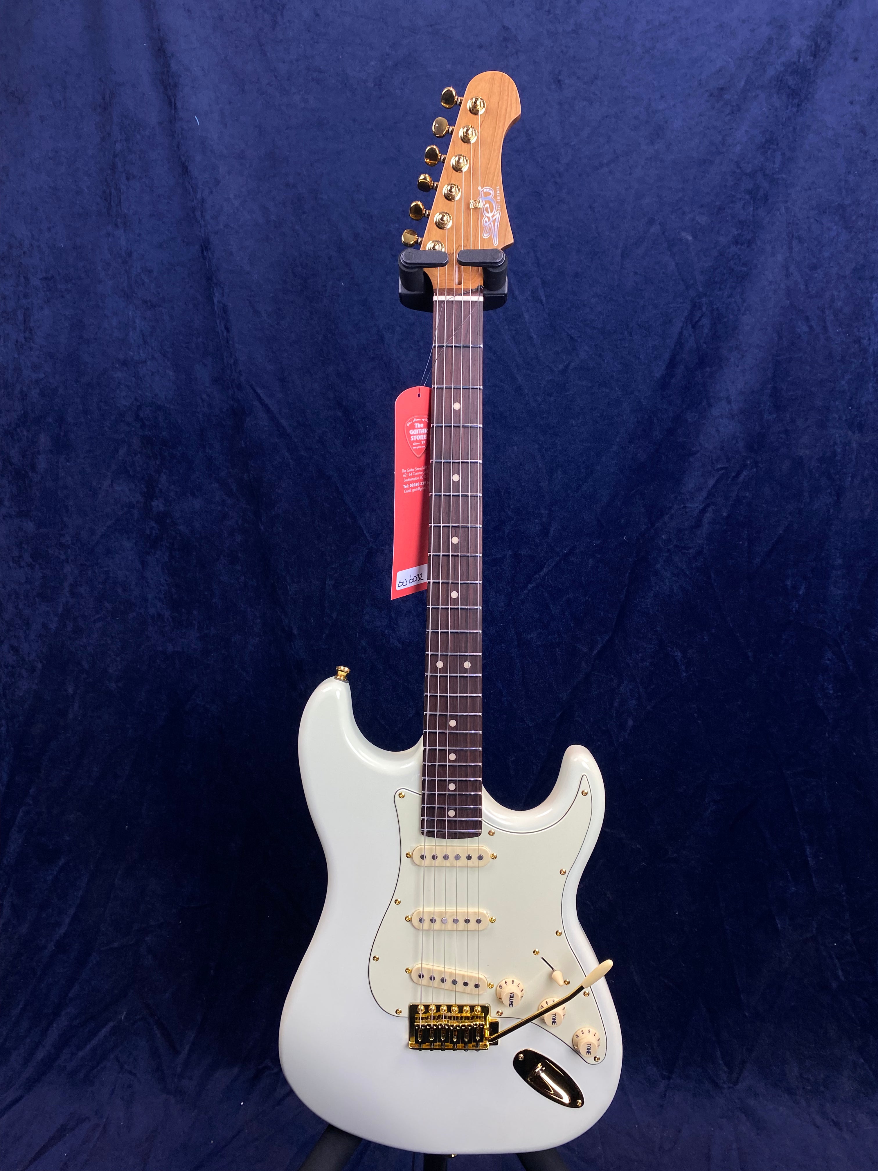 White electric guitar with three single-coil pickups, gold hardware, dark fretboard, and wooden headstock, displayed against a dark blue background.
