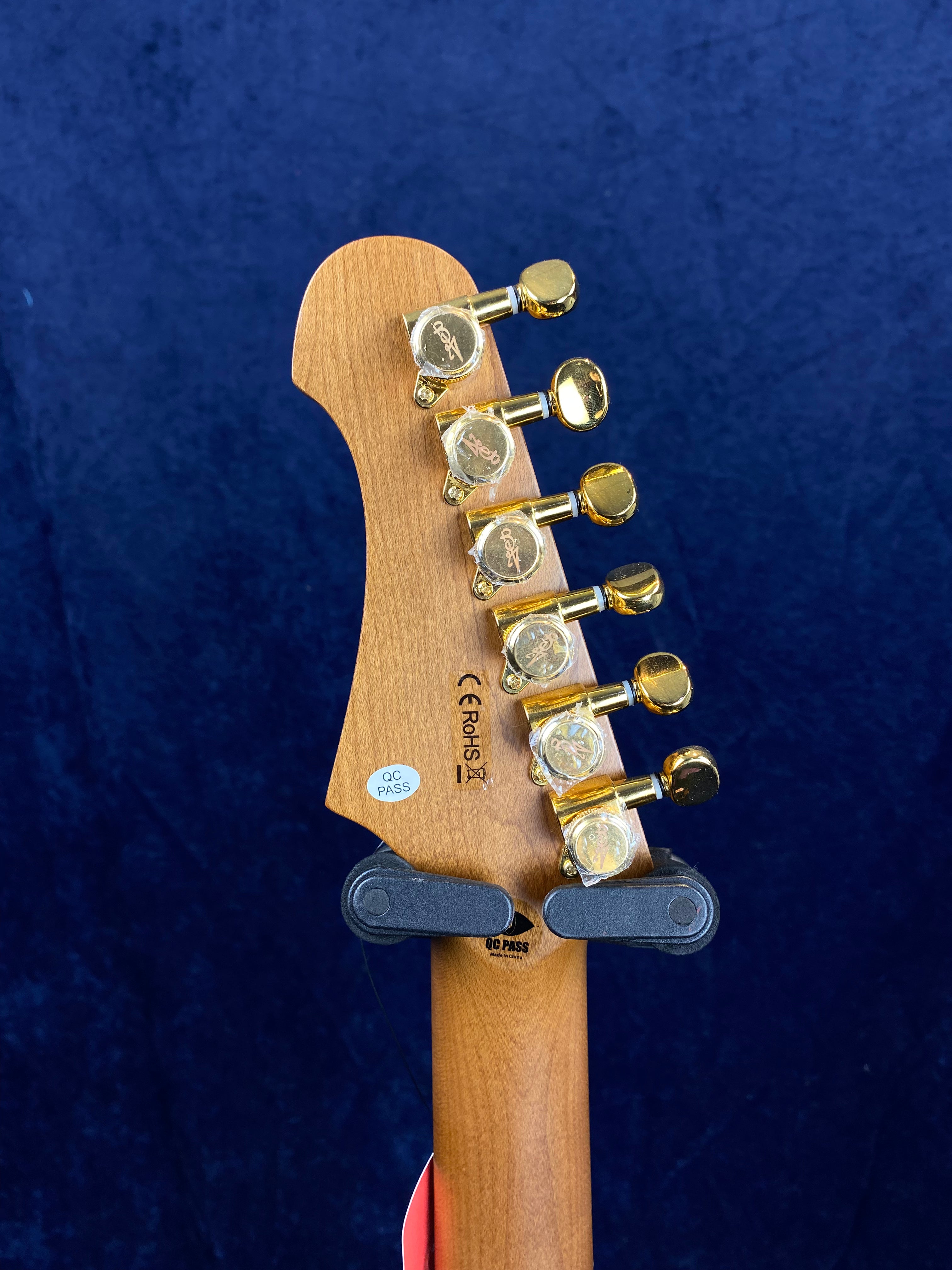 Six gold tuning pegs on the headstock of an electric guitar, light wood finish, quality control and certification stickers visible.