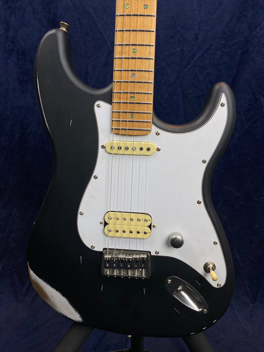 Black electric guitar with a white pickguard, maple neck, two single-coil pickups, one humbucker, and visible wear on the lower left body edge.