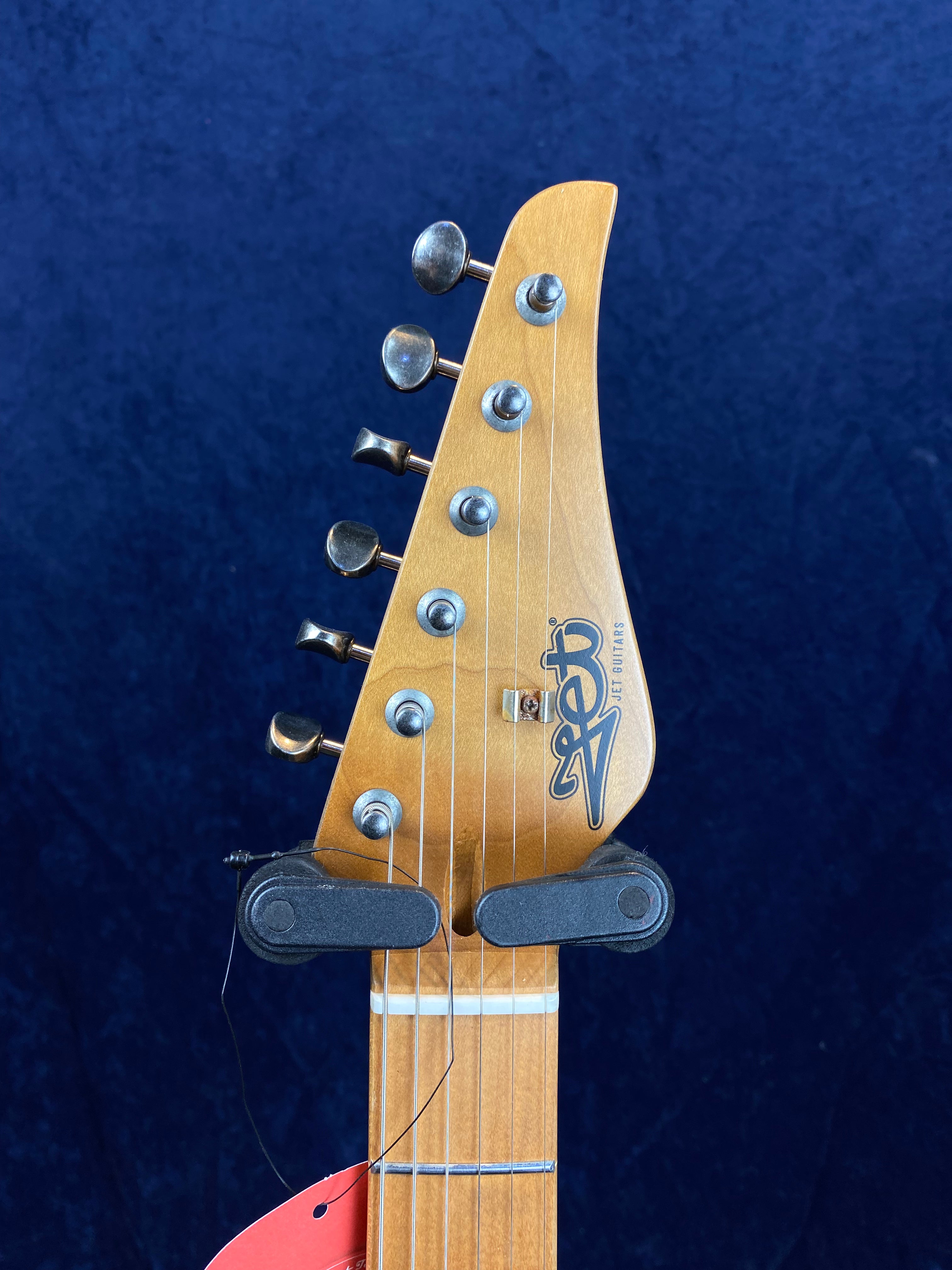 Electric guitar headstock with six tuning pegs, logo reading “Jet Guitars,” and a capo attached to the neck. Blue background.