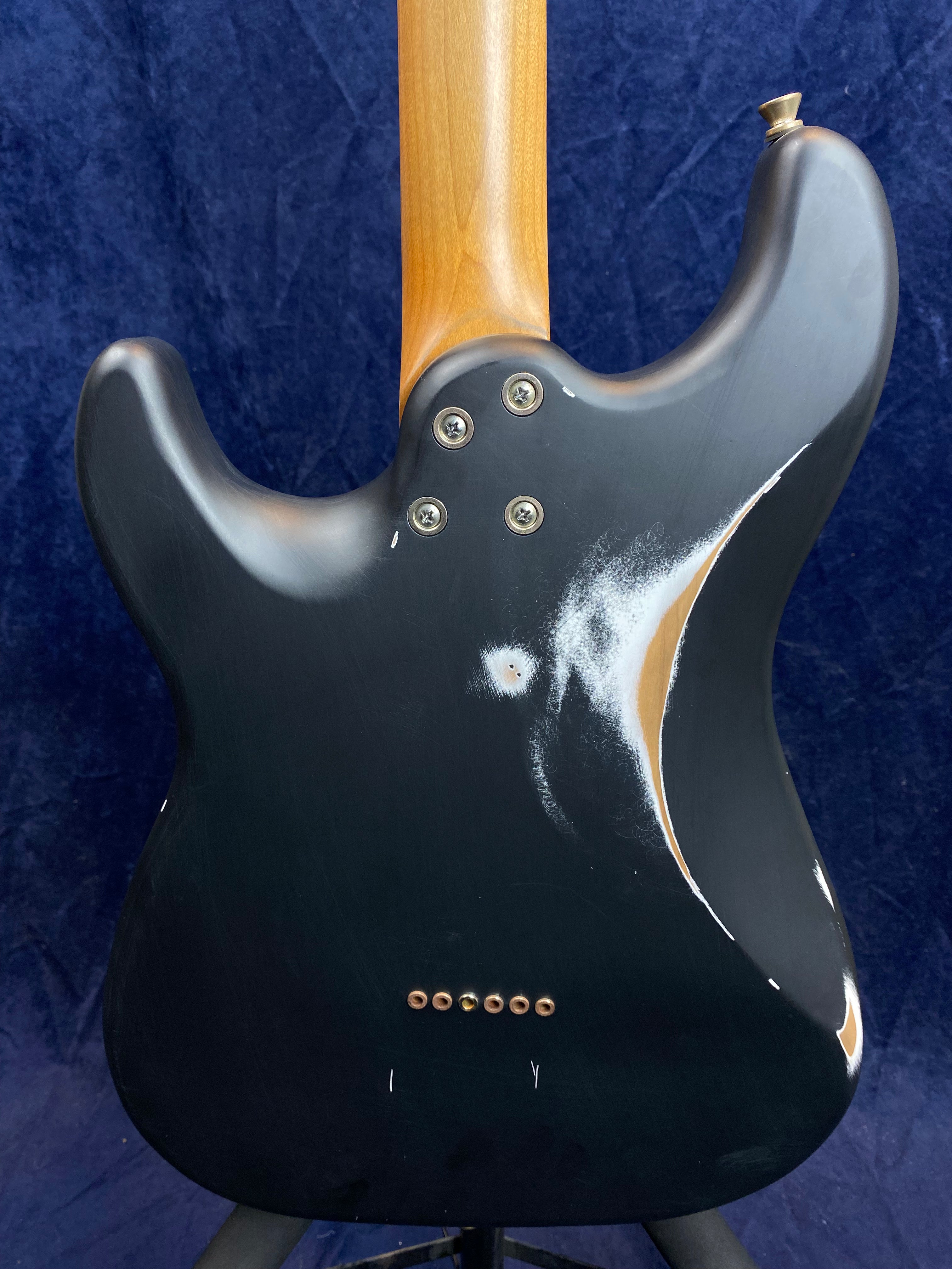 Back of an electric guitar with a matte black finish, visible scratches, worn spots revealing wood, and six string ferrules near the bottom.