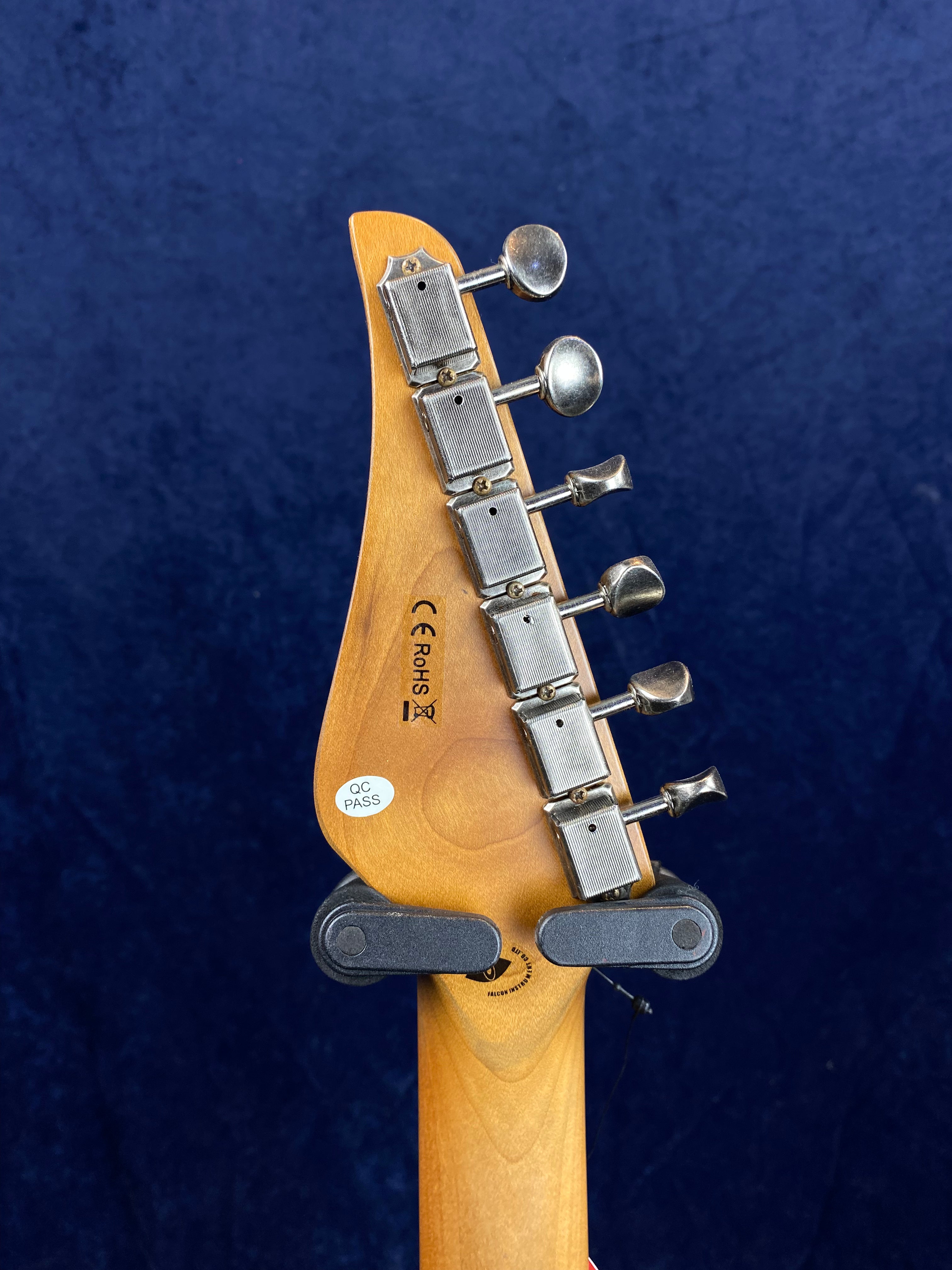 Back of an electric guitar headstock showing six vintage-style tuning pegs, light wood finish, QC PASS sticker, and part of a wall hanger.